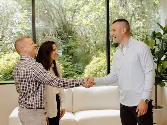 Two men shaking hands and laughing in a bright room with a woman smiling nearby.