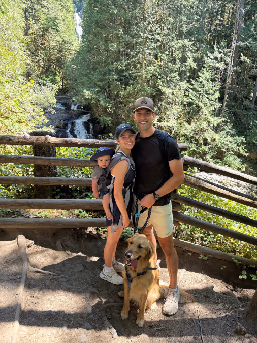 Family of three with a golden retriever posing on a wooded hiking trail near a waterfall.