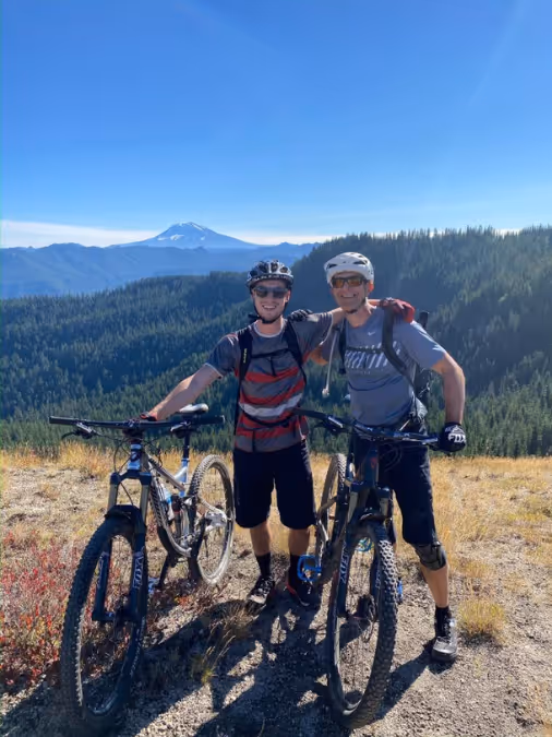 Two mountain bikers posing with bikes on a trail with forested hills and a snow-capped mountain behind.