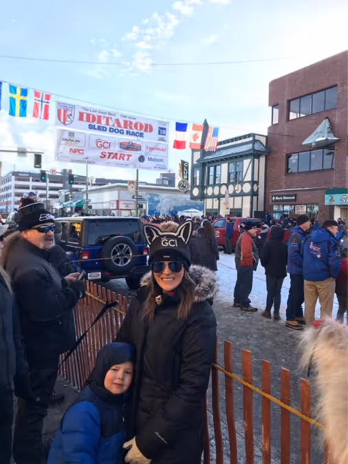 People in winter clothing gathered near the start line of the Iditarod sled dog race in a town.