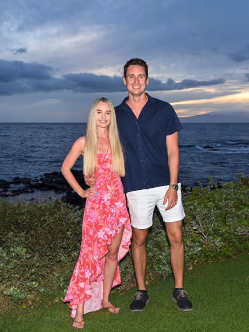 Couple standing by ocean at sunset; woman in pink floral dress, man in navy shirt and white shorts.