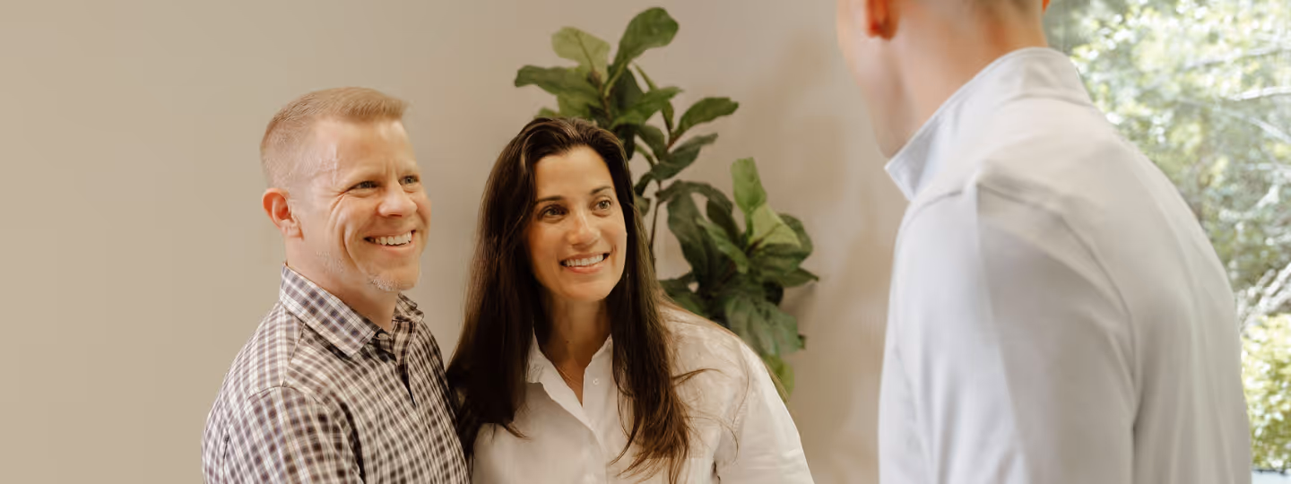 Smiling couple speaking with a man in a white shirt with a plant and window background.