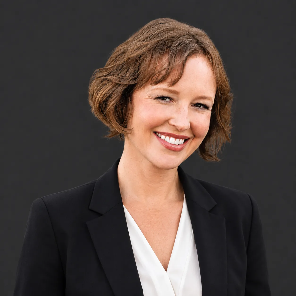 Professional headshot of Kristin Van Ausdal Senior Principal at MMC, a woman with shoulder-length dark brown hair wearing a dark top and a delicate necklace, smiling against a dark background.