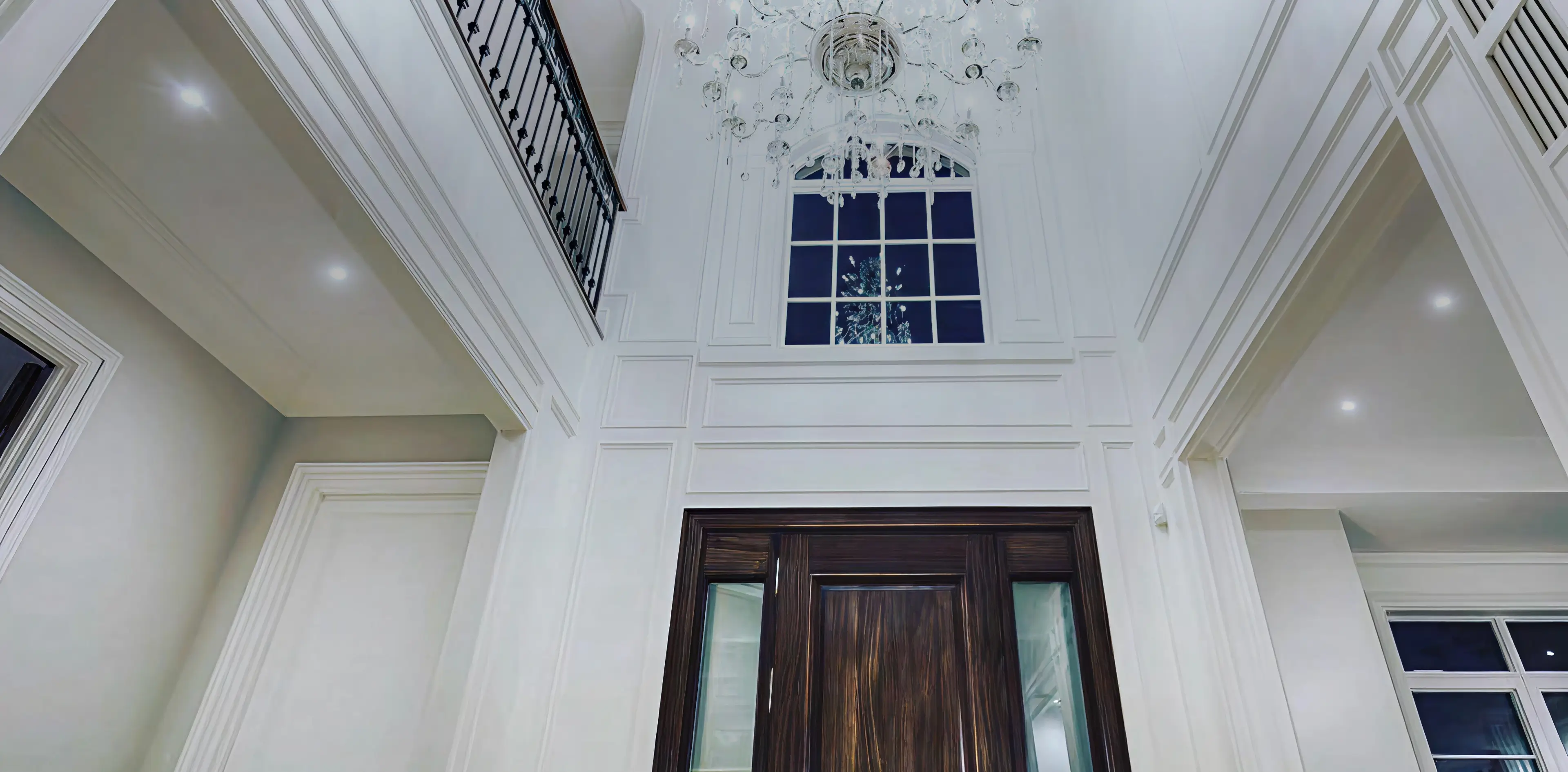 Interior view of a white-walled entryway with a dark wooden door, black metal railing balcony above, and a large chandelier.