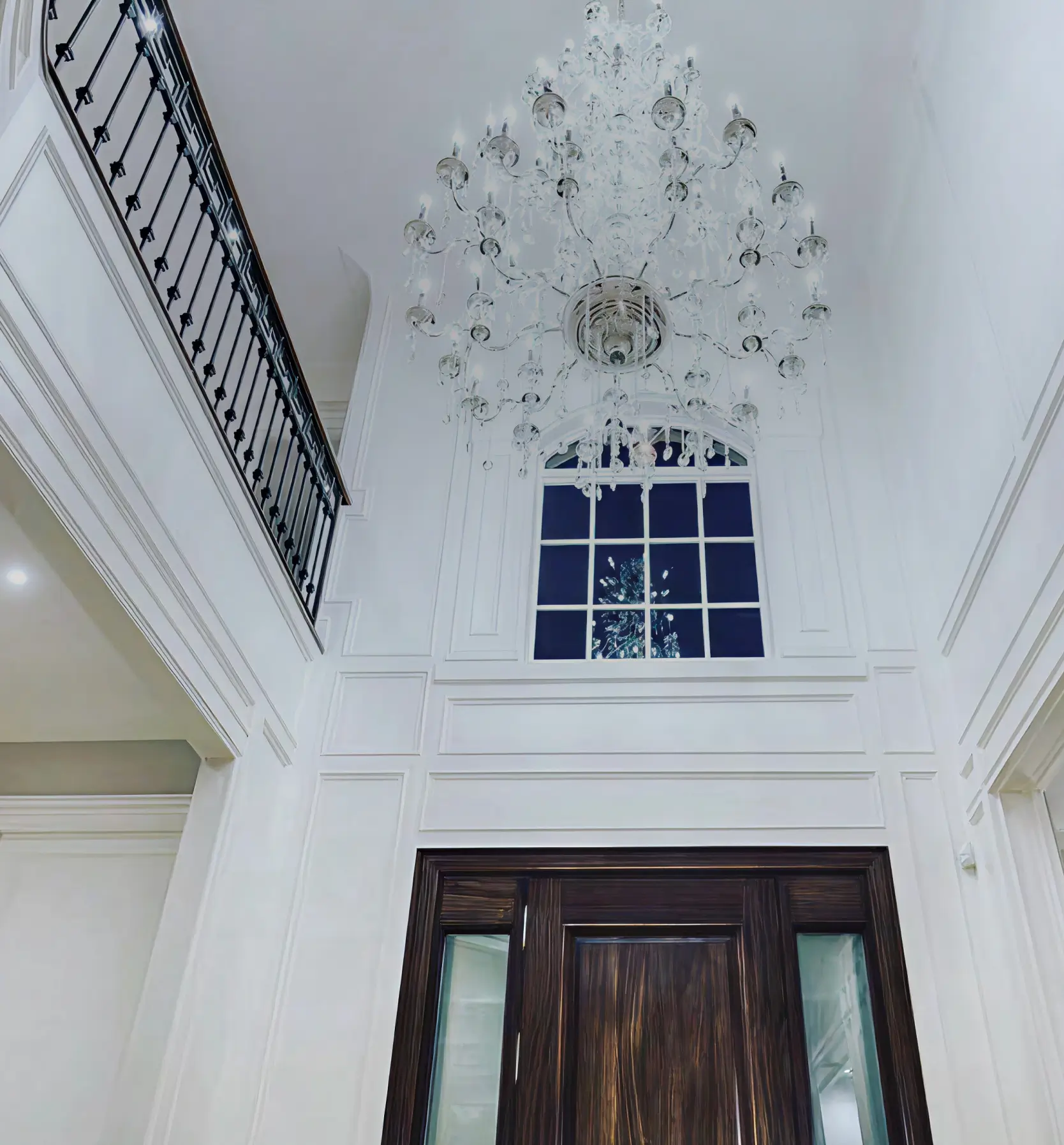 Interior view of a high-ceilinged room with white walls, a large ornate crystal chandelier, a tall window, and dark wooden double doors.