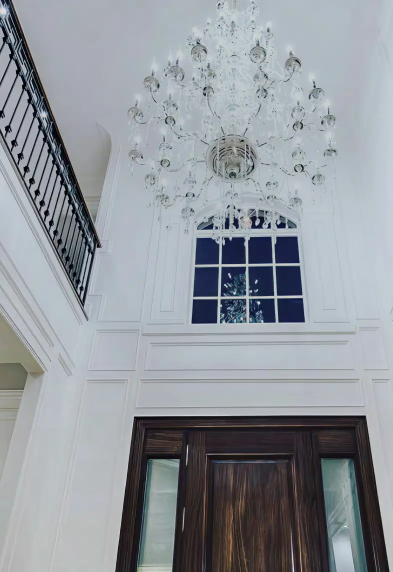 Large elegant crystal chandelier hanging in a white high-ceiling room above a dark wooden door with a window above it.
