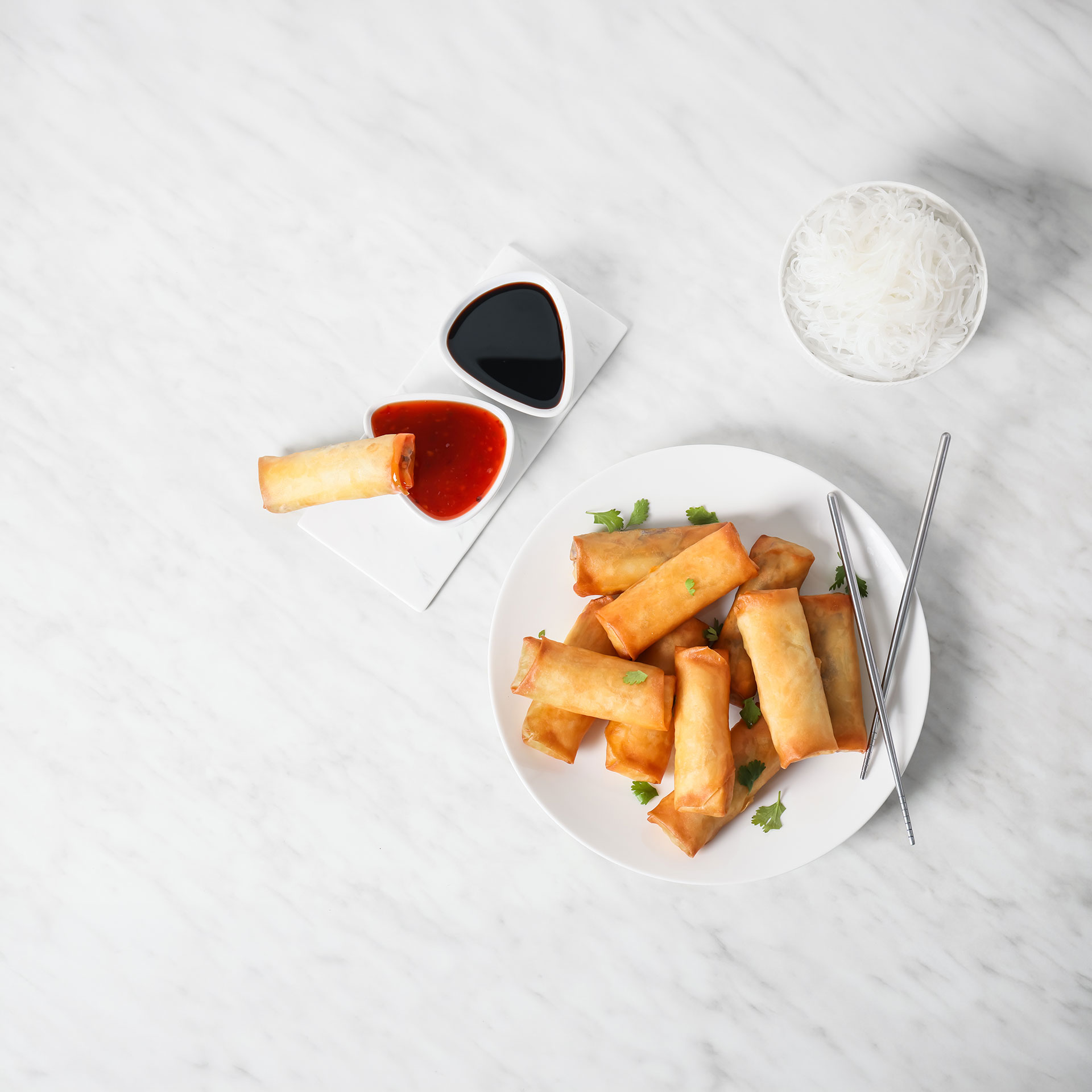 Plate of golden fried spring rolls garnished with cilantro, with two dipping sauces and a bowl of white rice noodles on a marble surface.