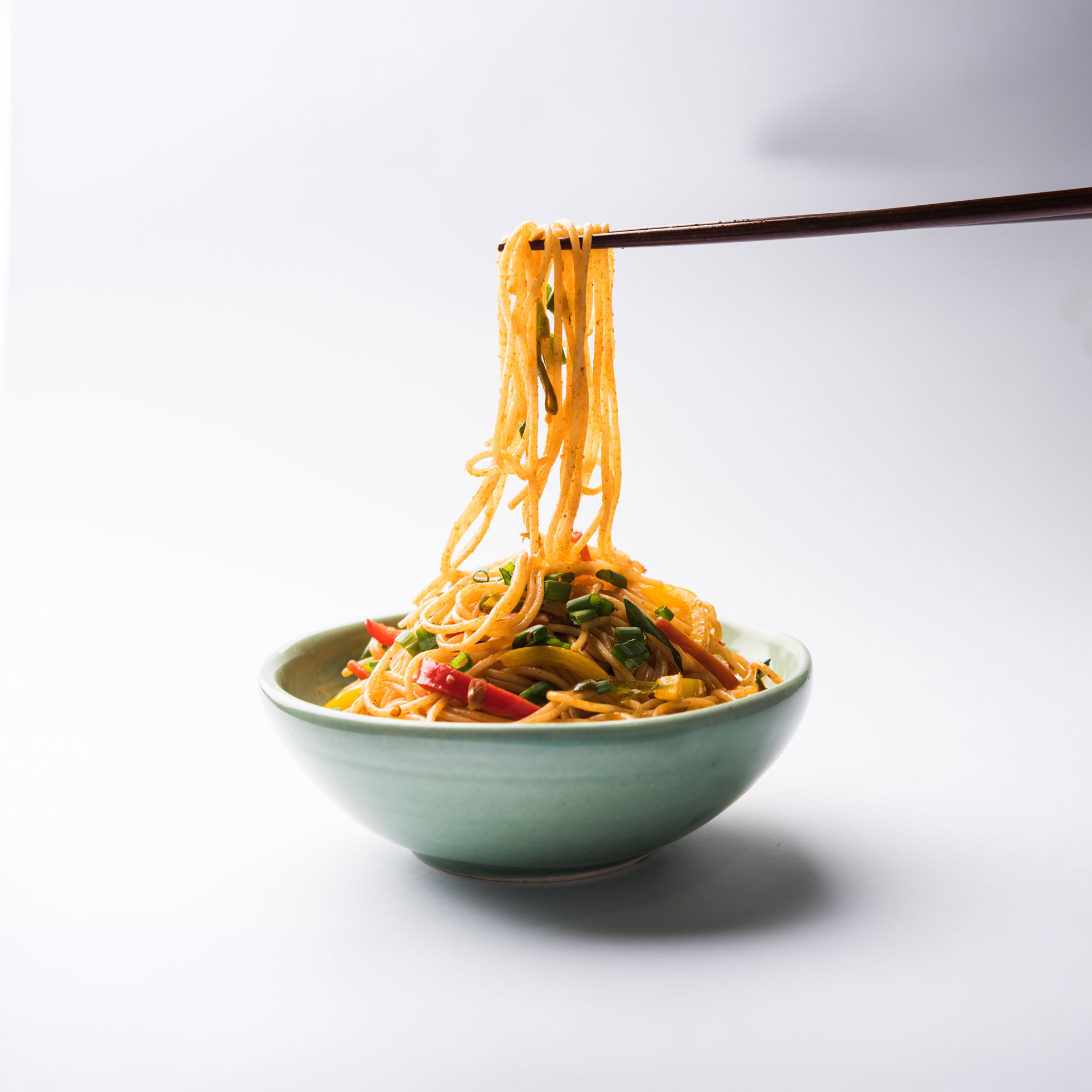 Chopsticks lifting seasoned noodles with vegetables from a green bowl on a white background.