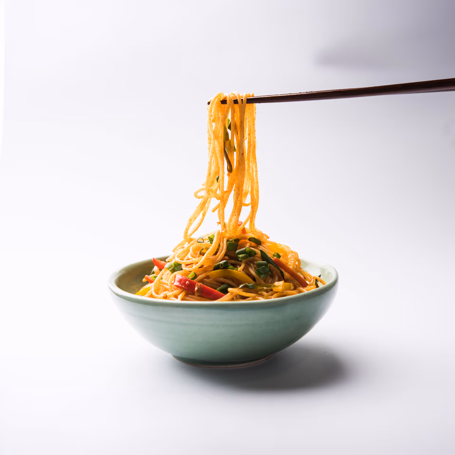 Chopsticks lifting seasoned noodles with vegetables from a green bowl on a white background.