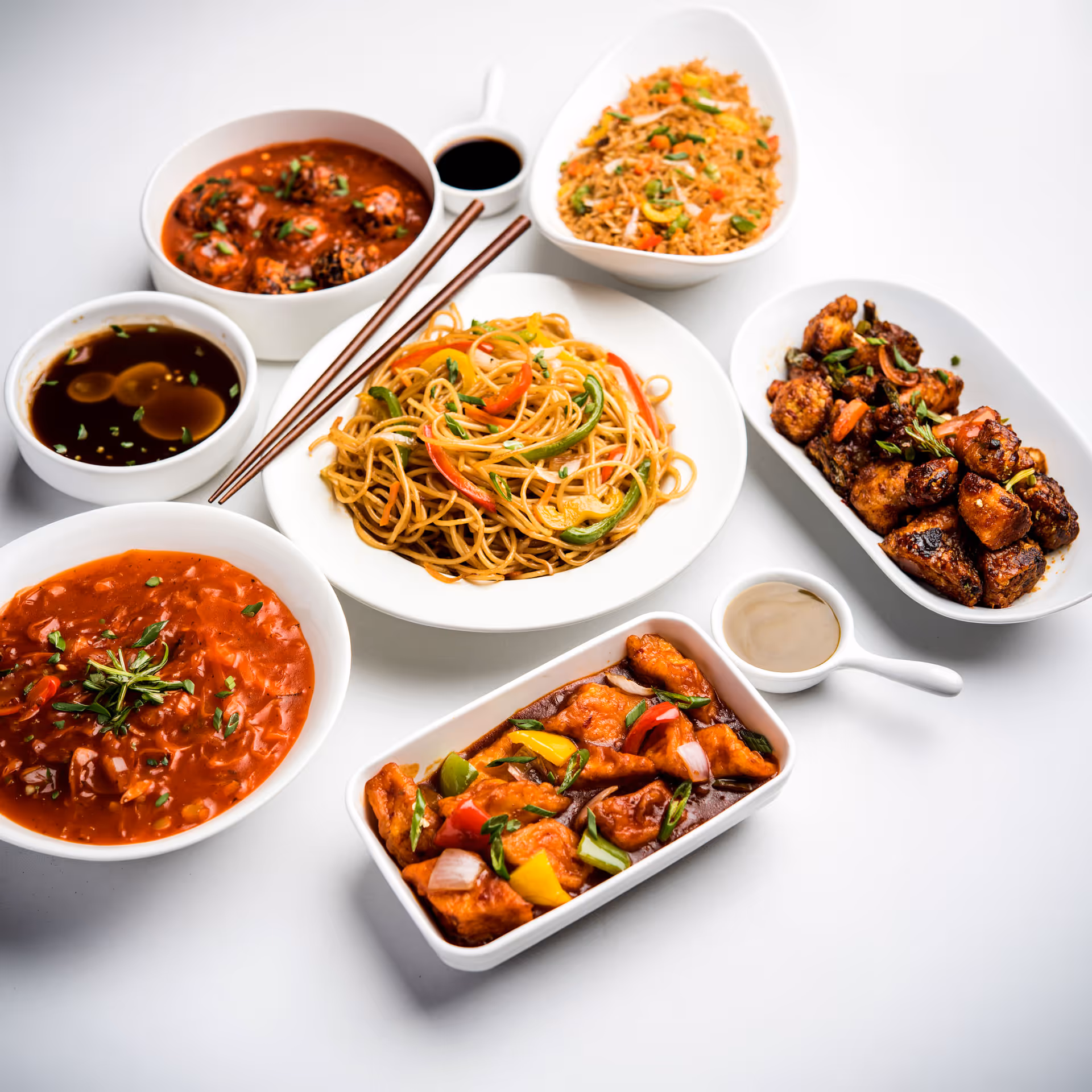 Variety of Chinese dishes including stir-fried noodles with bell peppers, fried rice, and several sauced vegetable and protein dishes with chopsticks on a white surface.