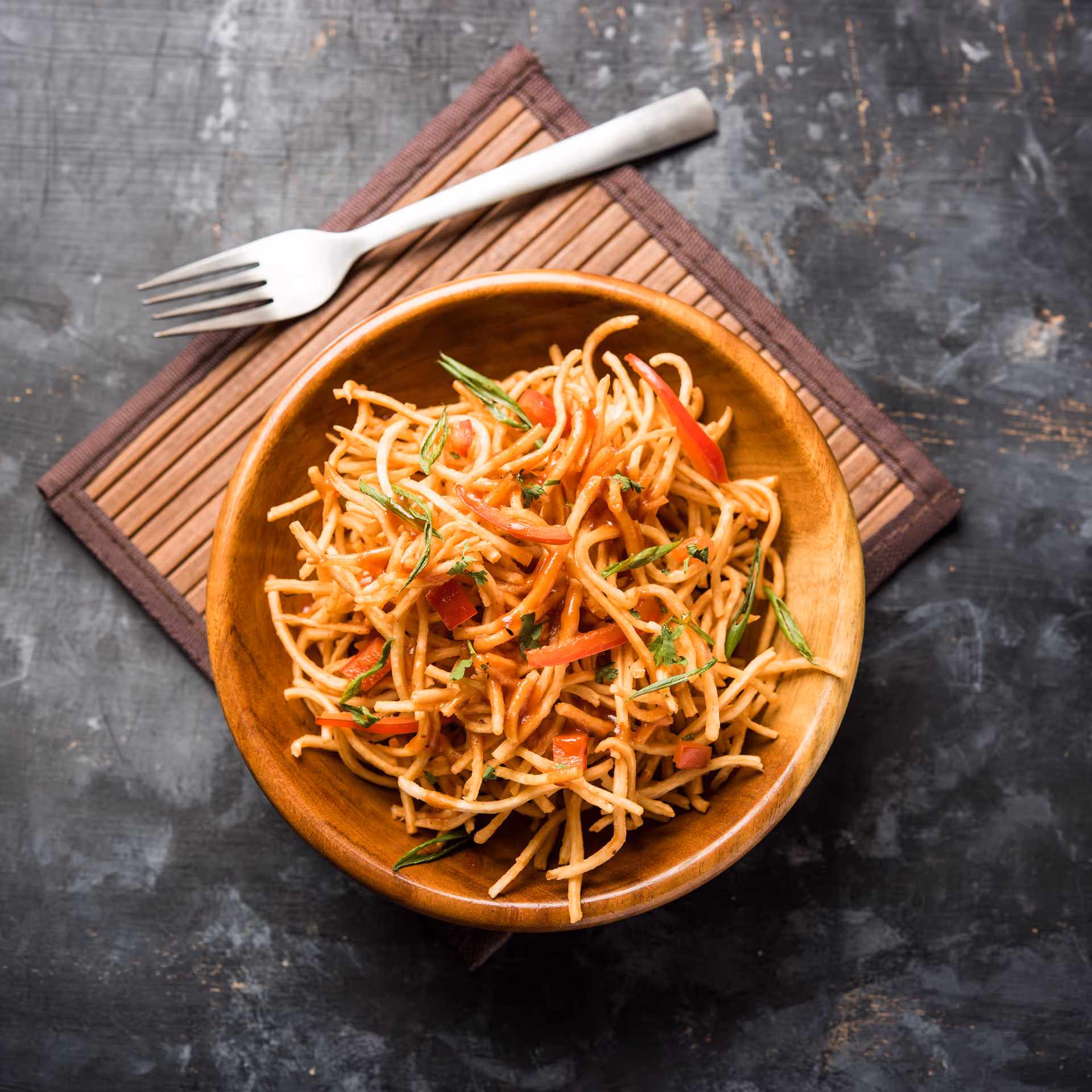 Bowl of crispy fried noodles garnished with sliced red bell peppers and green herbs on a bamboo mat with a fork on a dark textured surface.