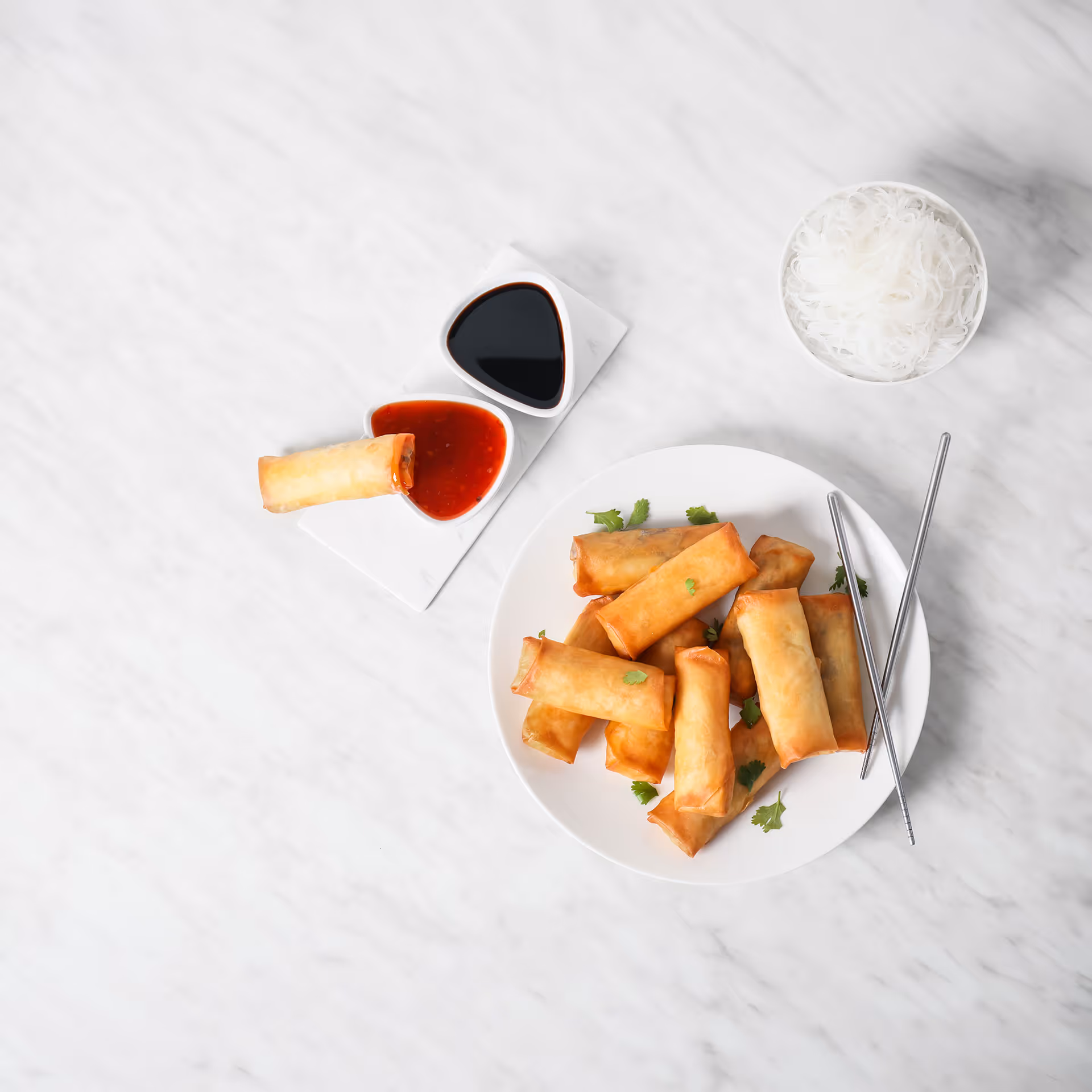 Plate of golden fried spring rolls garnished with cilantro, with two dipping sauces and a bowl of white rice noodles on a marble surface.
