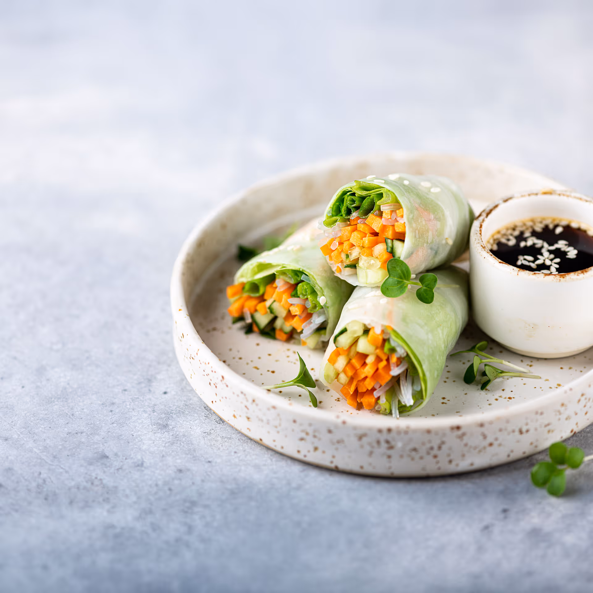 Three fresh vegetable spring rolls with carrots, cucumbers, and greens on a white speckled plate with a small cup of soy sauce topped with sesame seeds.