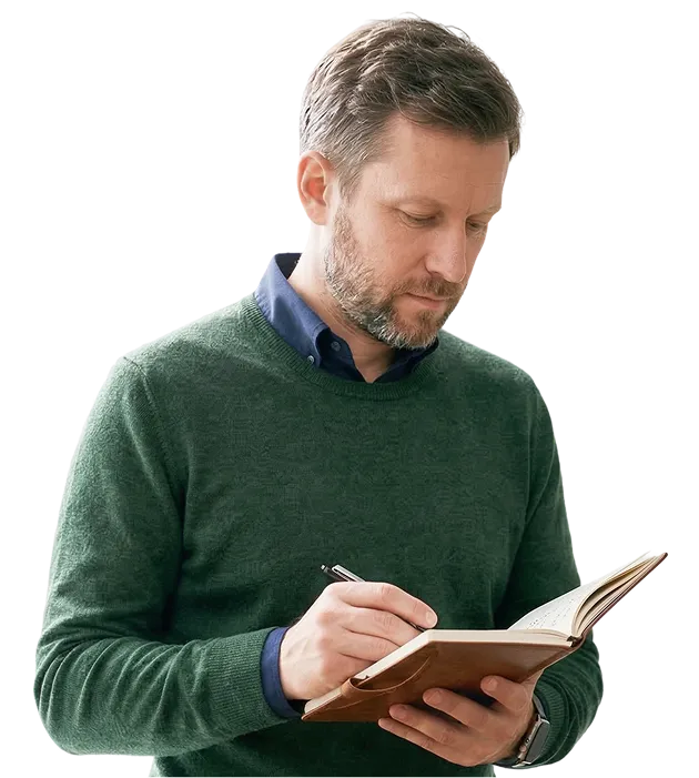 Man wearing a green sweater and blue shirt writing in a notebook.