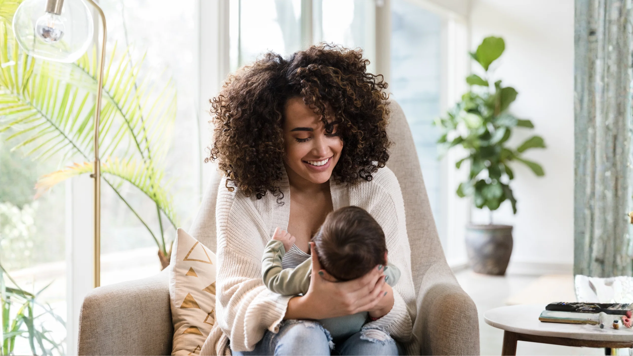 A smiling woman with voluminous curly hair tenderly cradles a baby while sitting in a bright, plant-filled living room.