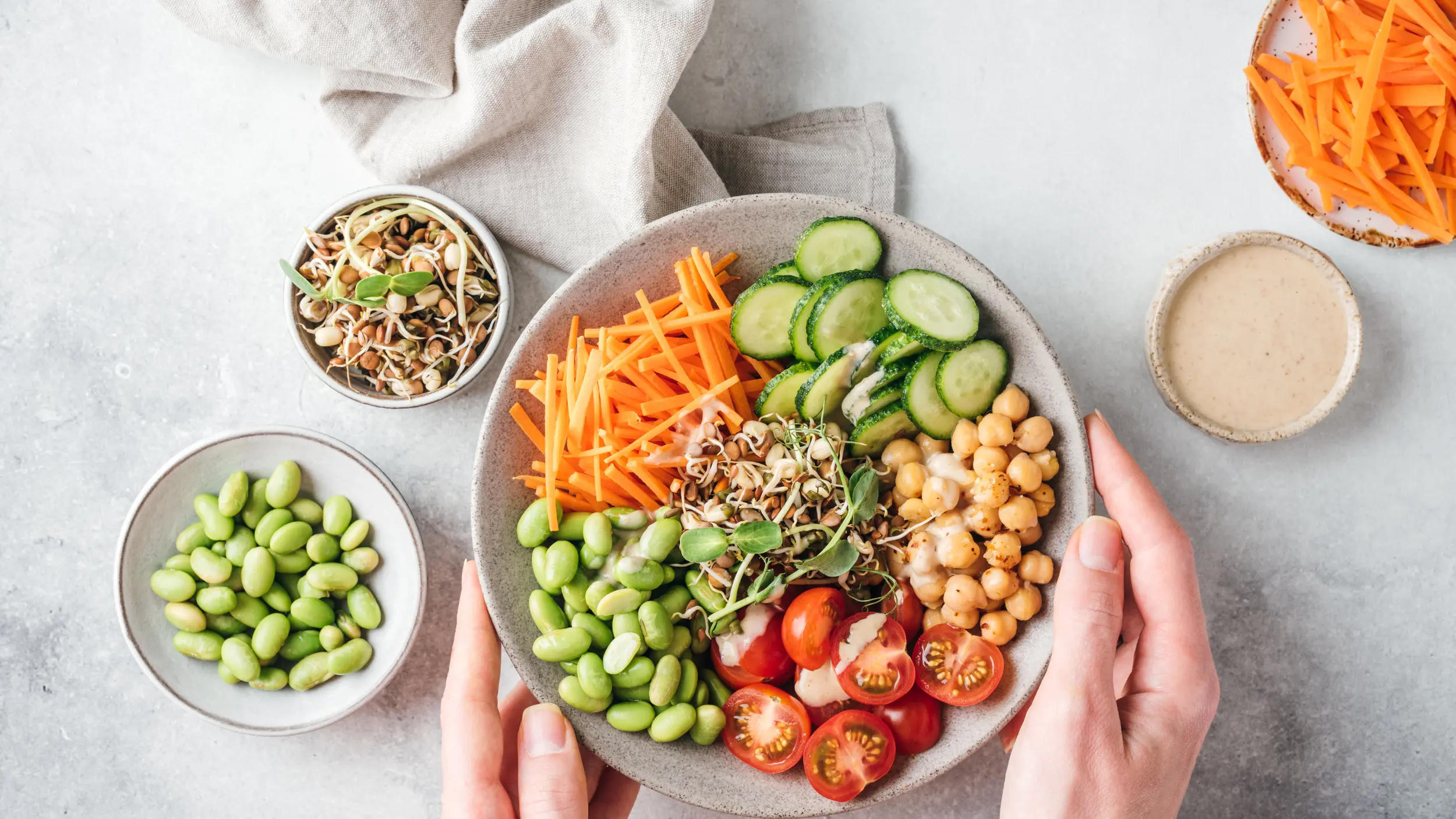 An overhead view of hands holding a colorful nourish bowl filled with sections of edamame, carrots, cucumbers, chickpeas, and cherry tomatoes, surrounded by small side dishes on a grey surface.