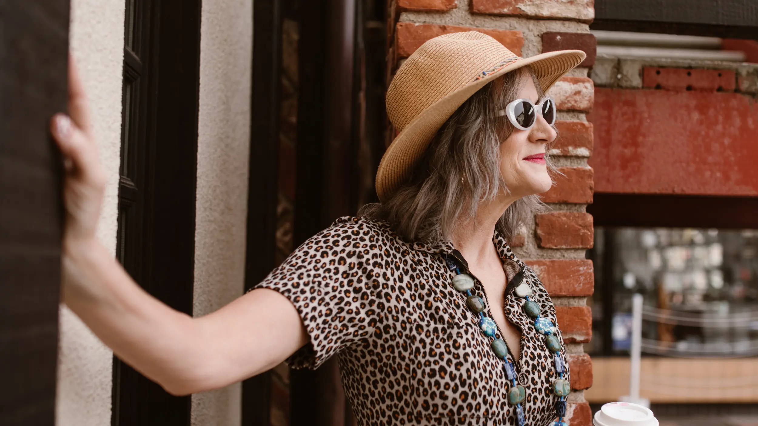 A stylish woman with grey hair wearing a straw hat, white-rimmed sunglasses, and a leopard-print top smiles while standing outdoors next to a brick wall.