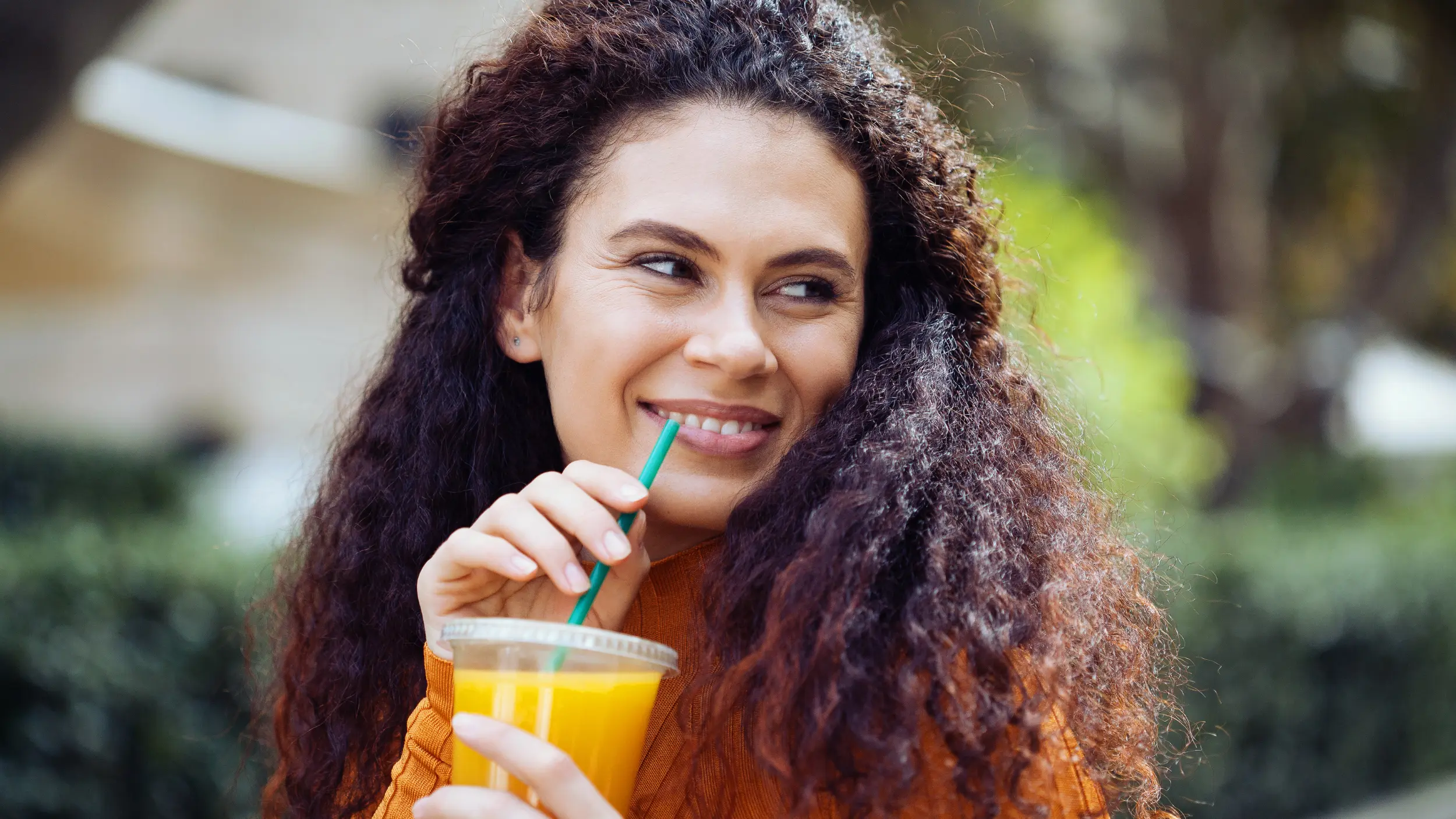 A smiling woman with long, curly hair drinks a vibrant orange adrenal support mocktail from a clear cup with a green straw outdoors.