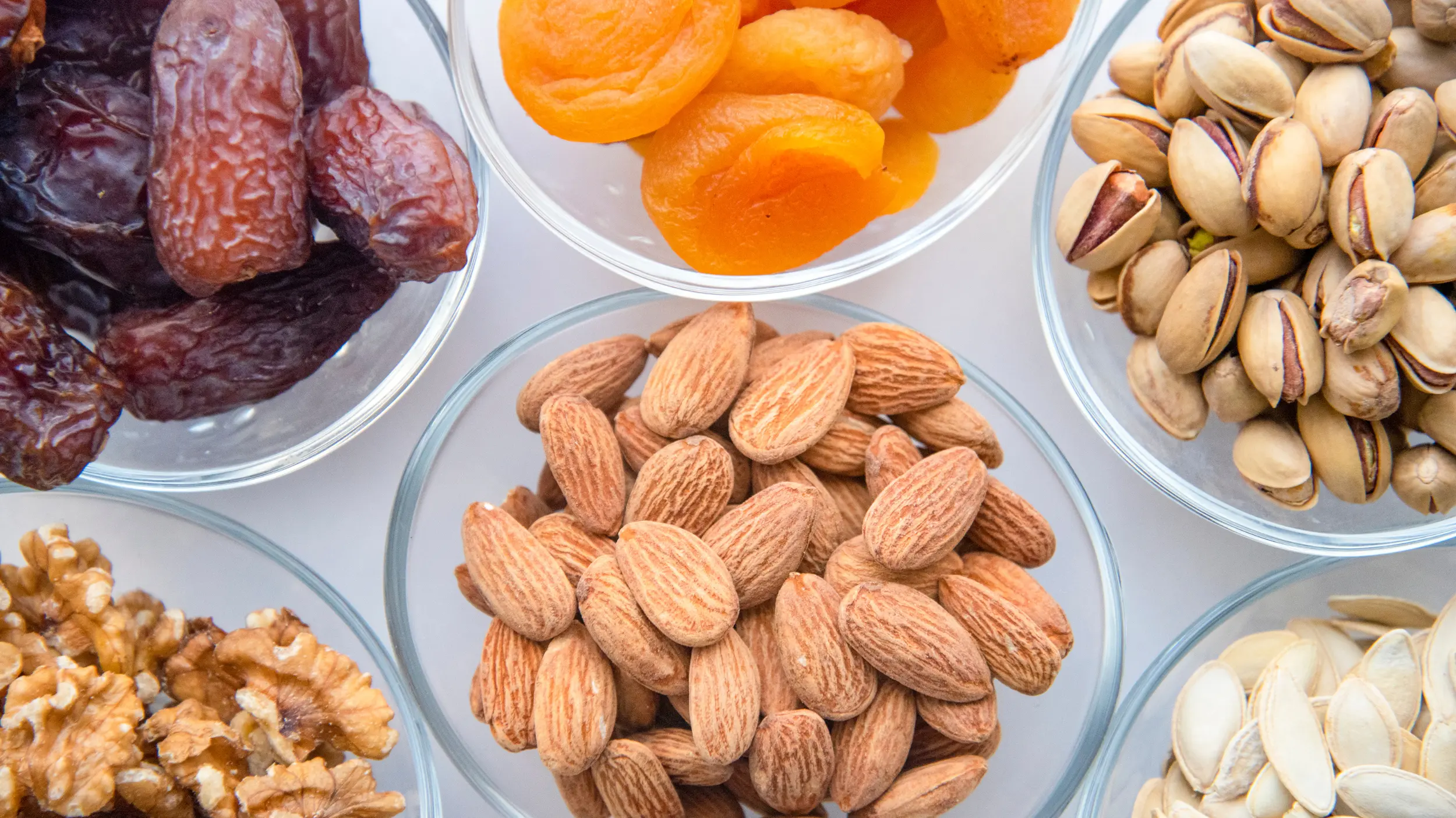 An overhead view of small glass bowls filled with a variety of healthy snacks, including almonds, dried apricots, pistachios, dates, walnuts, and pumpkin seeds.