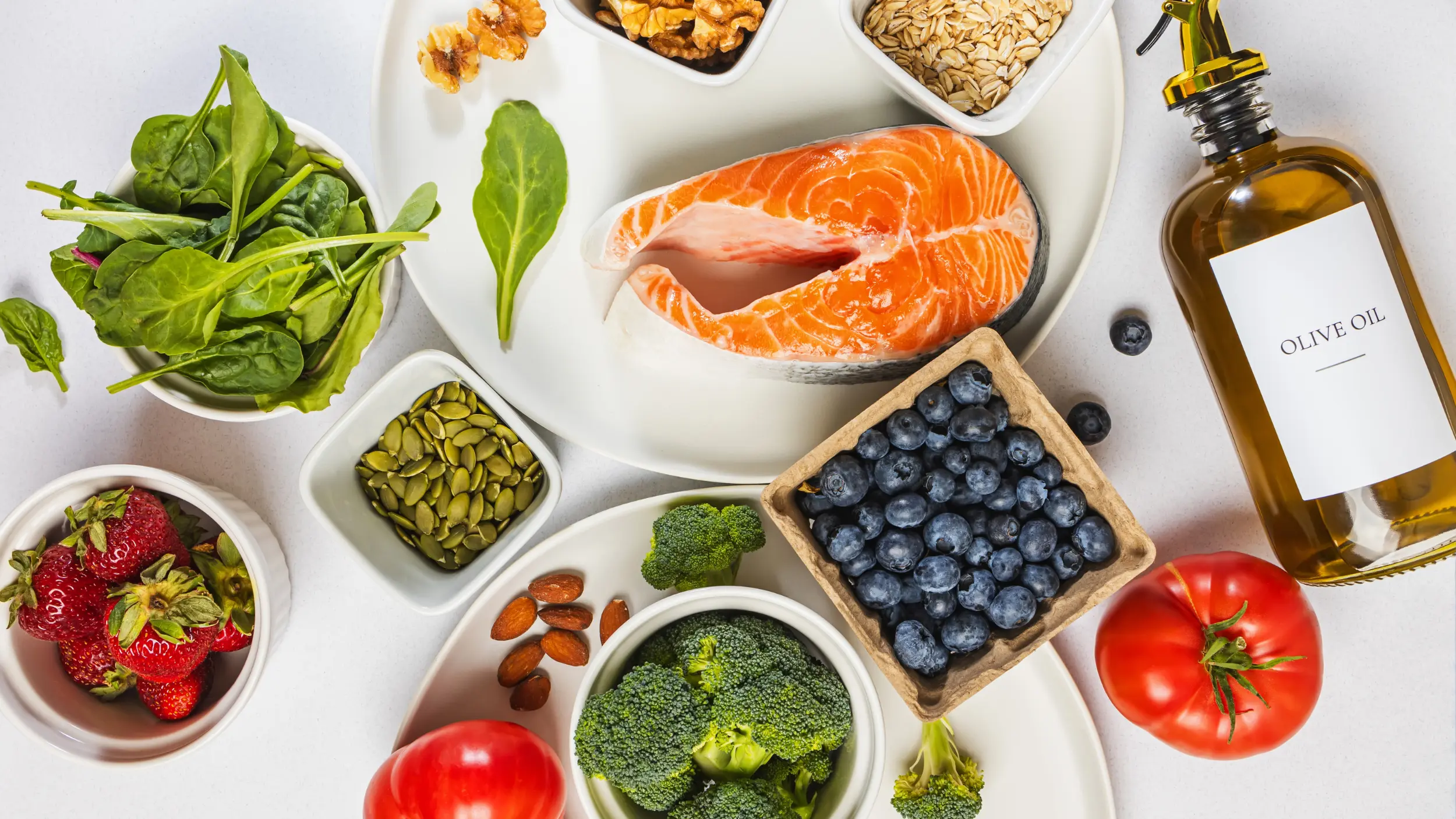 An overhead view of a selection of healthy foods, including a raw salmon steak, a bottle of olive oil, broccoli, tomatoes, blueberries, strawberries, spinach, nuts, and oats on a light surface.