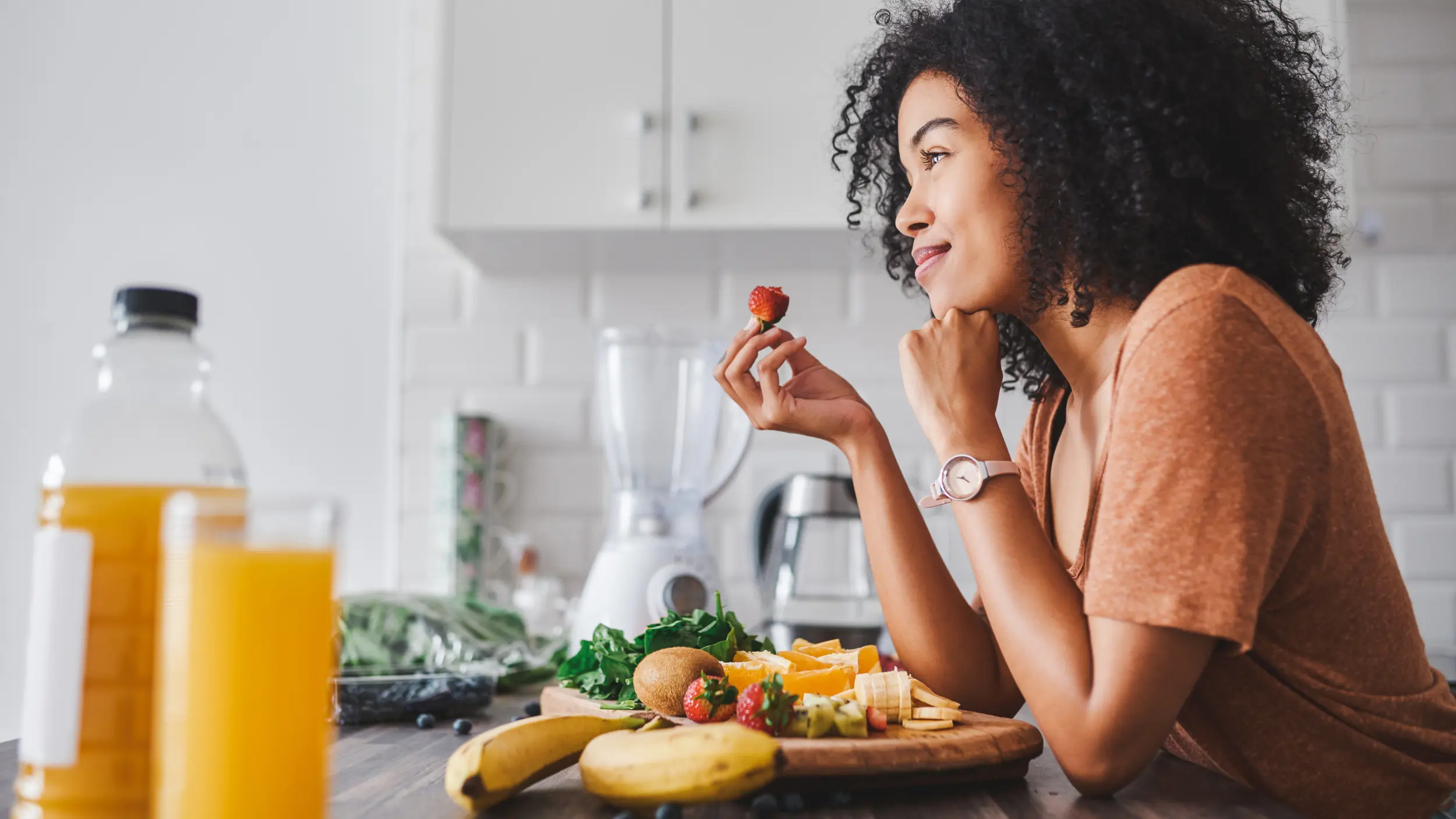 A woman with curly hair smiling thoughtfully while practicing intuitive eating, holding a strawberry at a kitchen counter filled with fresh fruits and a glass of orange juice.
