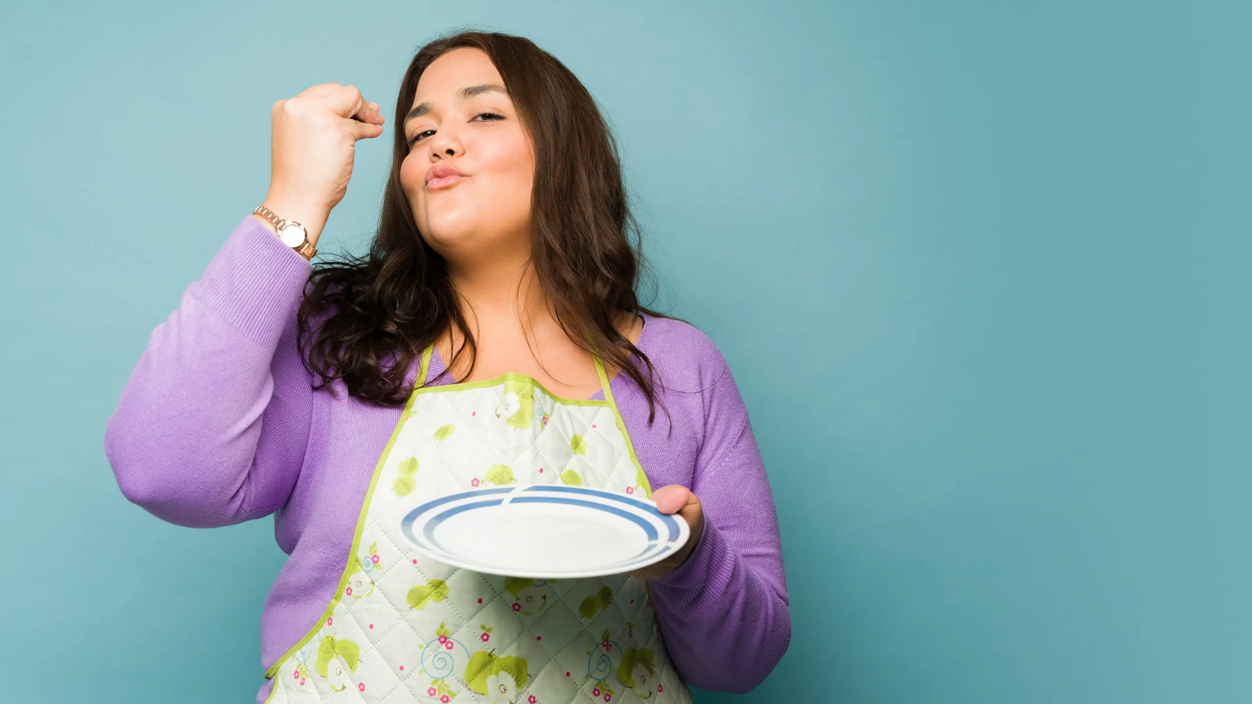 A woman wearing a purple sweater and an apron holds an empty plate while making a "chef's kiss" gesture against a solid teal background.