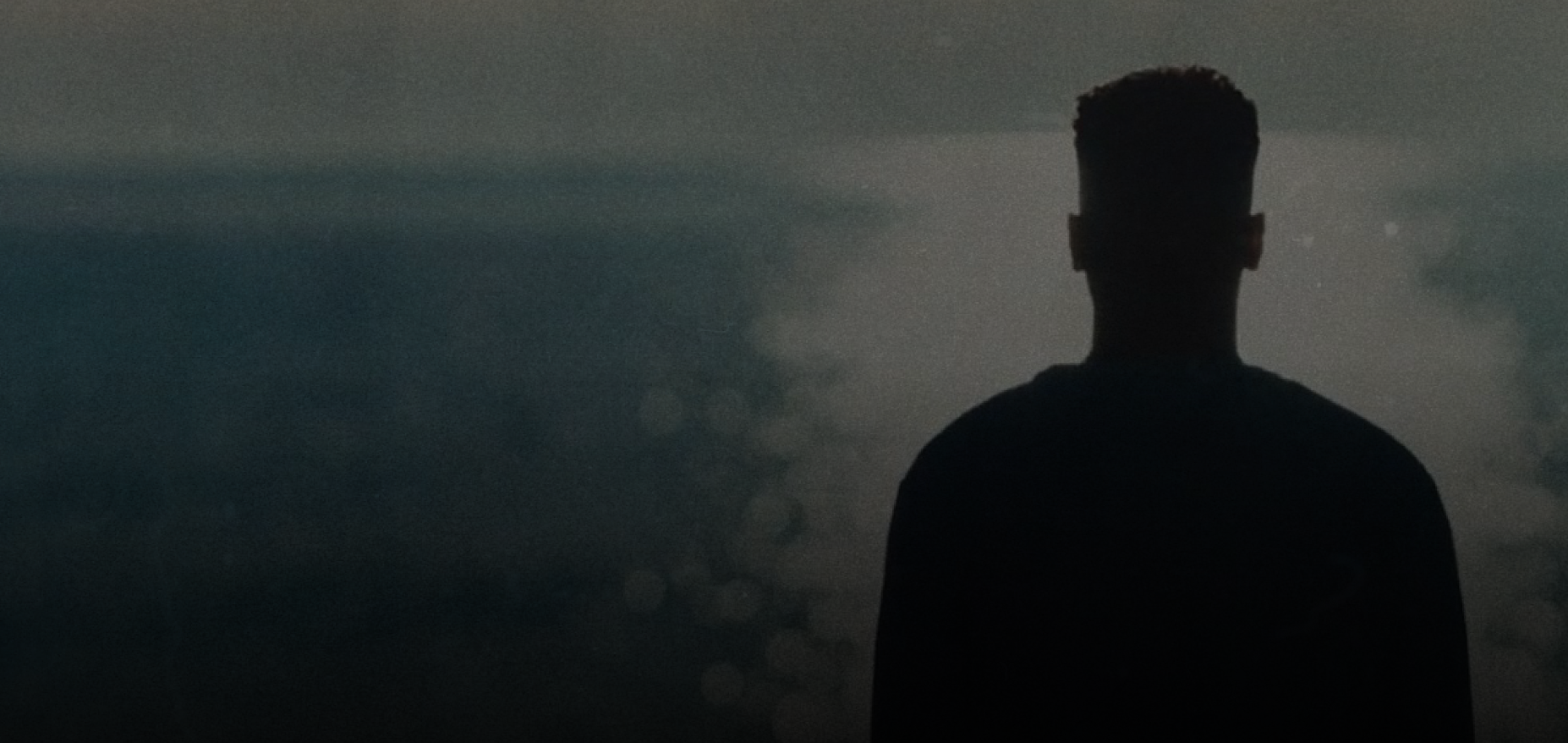 Silhouette of a man with short hair standing and facing a misty, dark background.