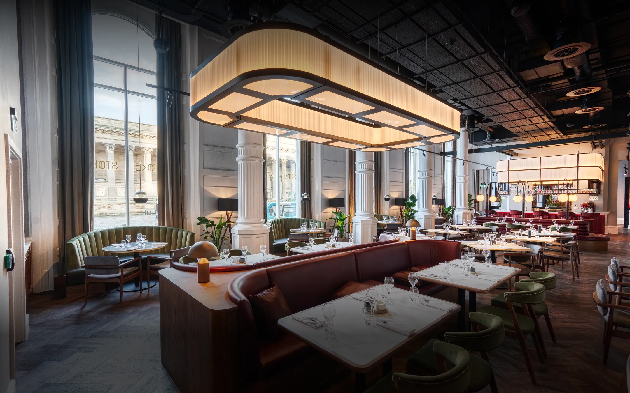 Elegant restaurant interior with curved booths, green chairs, large windows, and warm hanging lights.