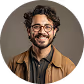 Smiling man with curly hair, glasses, and beard wearing a brown jacket and dark shirt.