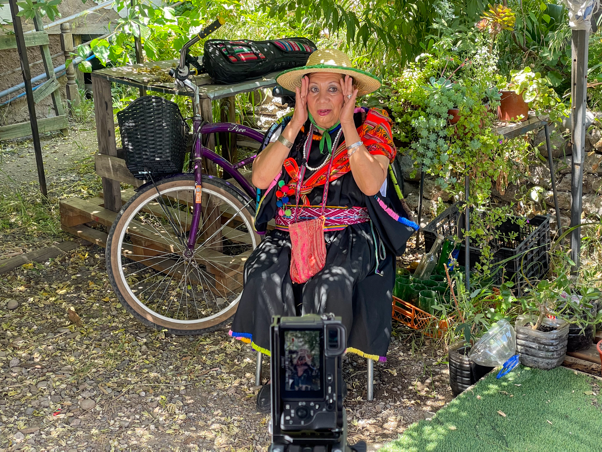 Mujer con vestimenta tradicional latinoamericana siendo grabada en jardín con bicicleta y plantas