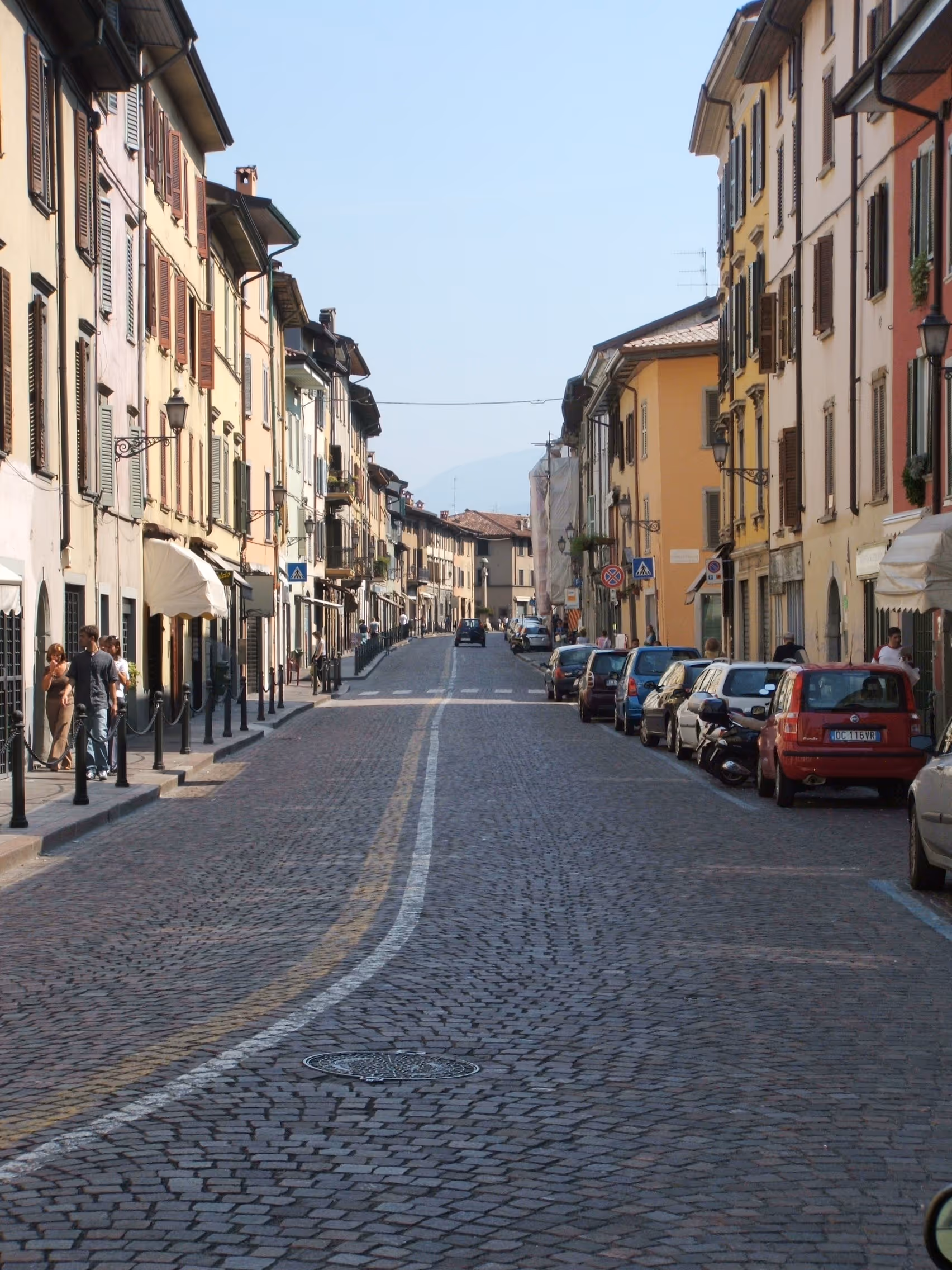 Strada acciottolata con auto parcheggiate e persone vicino a edifici colorati a Bergamo, Italia.
