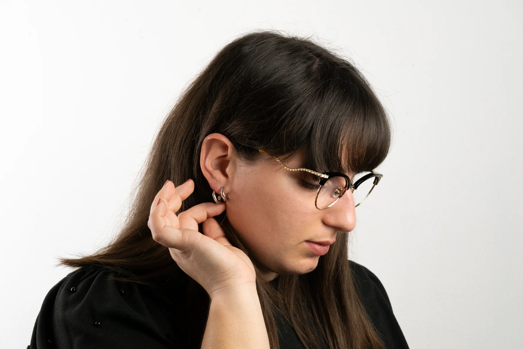 Ragazza con occhiali neri e oro che tocca una orecchino a cerchio argento, vista laterale con sfondo bianco.
