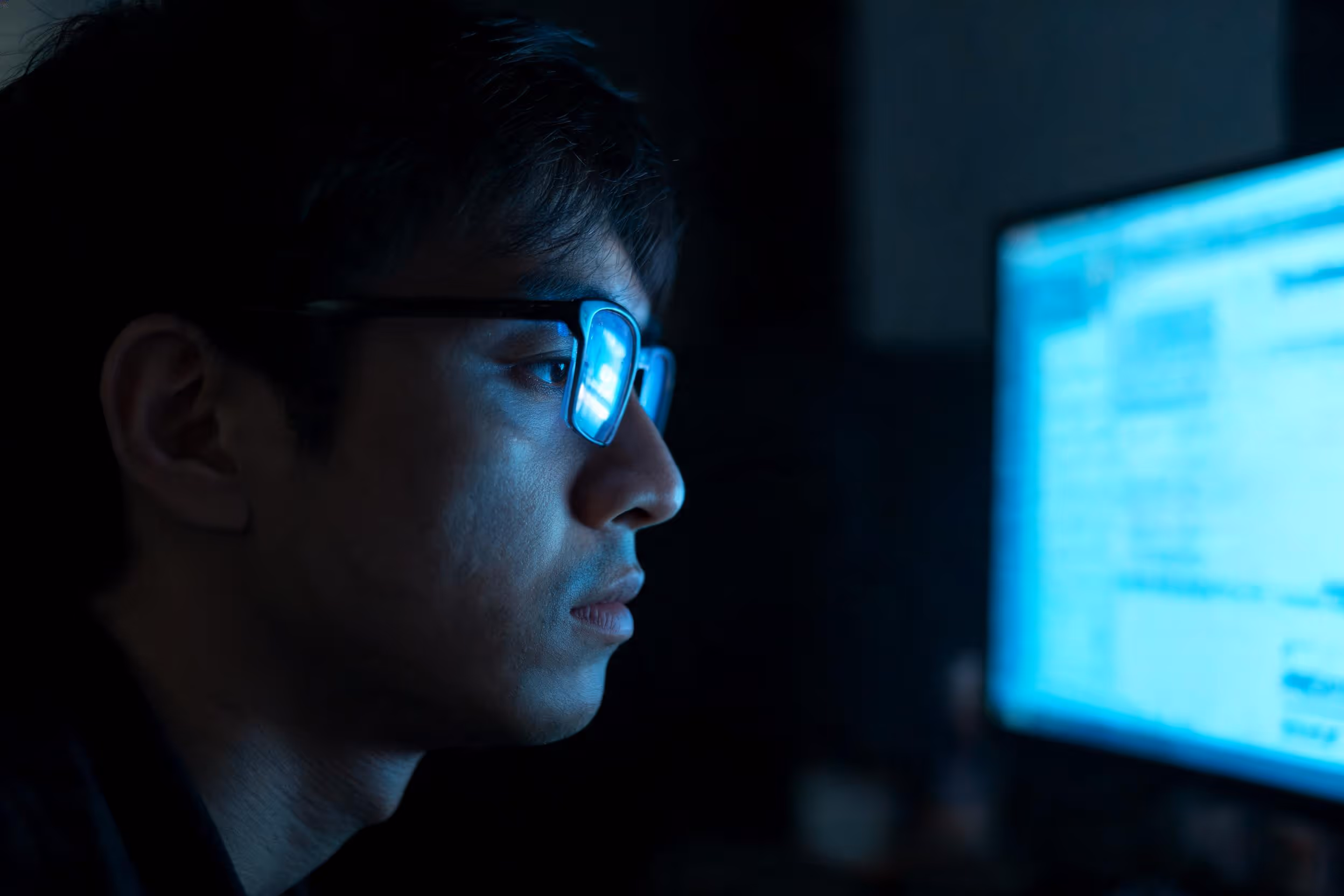 Uomo con occhiali guarda uno schermo illuminato al computer in una stanza buia.