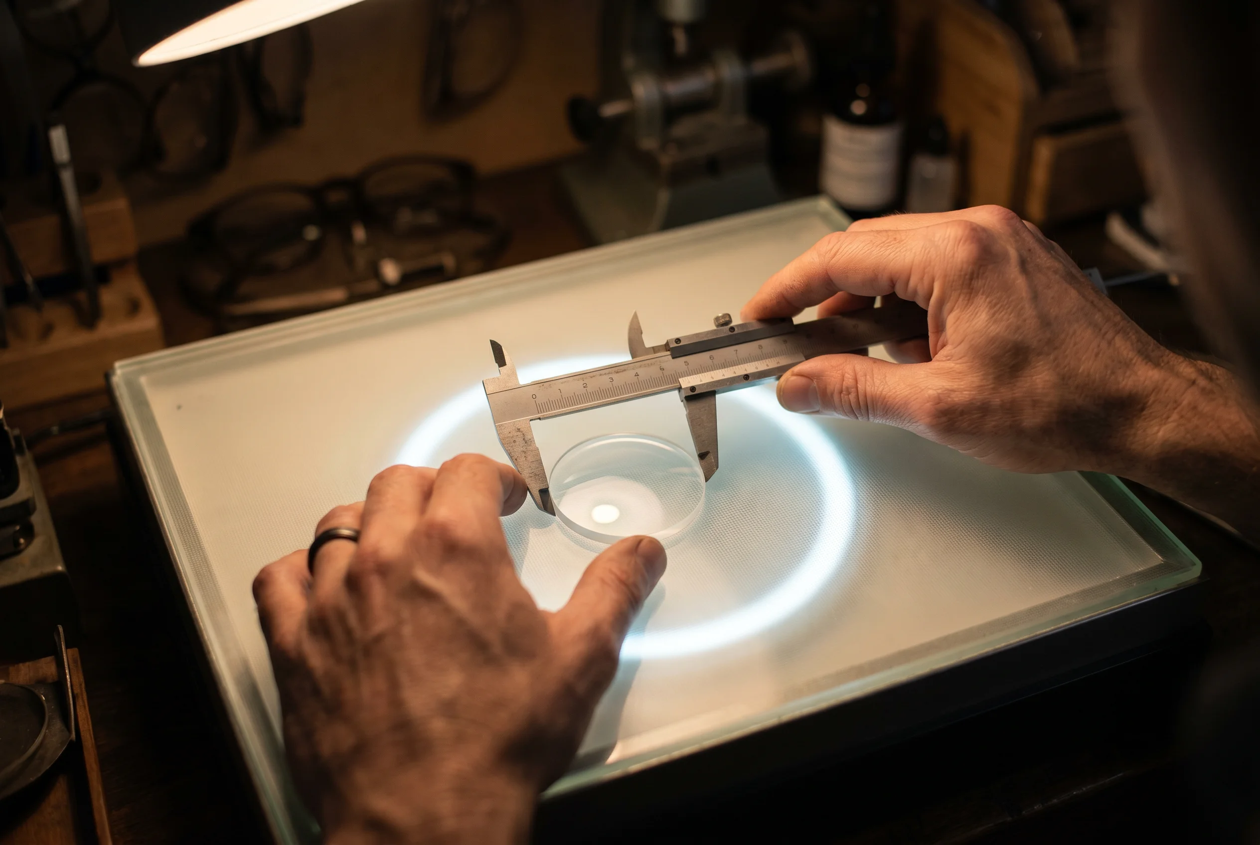 Man using caliper to measure diameter of a round glass lens on a lit table surface.