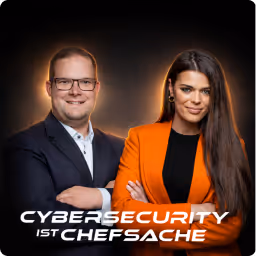 Porträt von einem Mann mit Brille in dunkelblauem Anzug und einer Frau mit langem Haar und oranger Jacke vor dunklem Hintergrund.