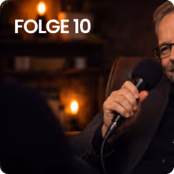 Ein Mann mit Brille hält ein Mikrofon in einem dunklen Raum mit warmem Licht.