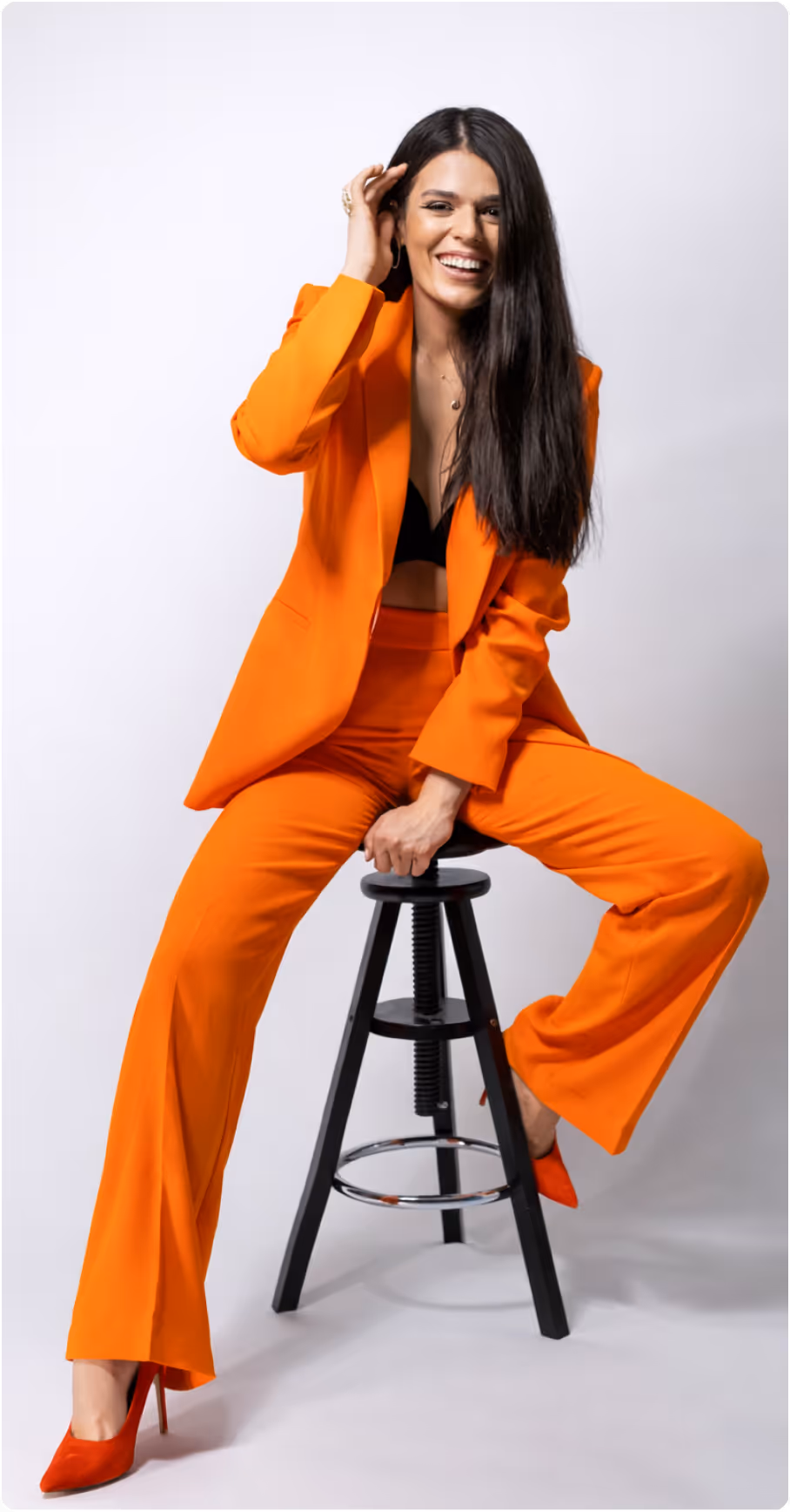 Frau mit langen dunklen Haaren trägt einen orangefarbenen Hosenanzug und rote Pumps, sitzt lachend auf einem schwarzen Hocker.