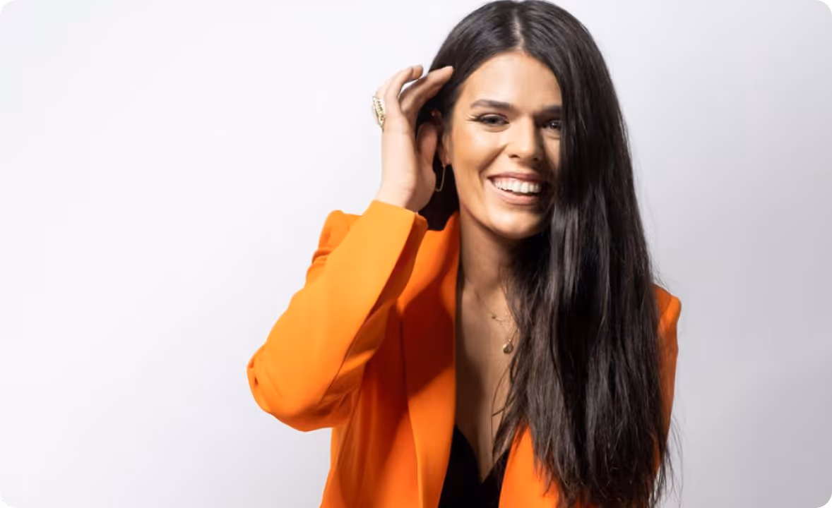 Lächelnde Frau mit langem dunklem Haar und orangefarbenem Blazer vor weißem Hintergrund.