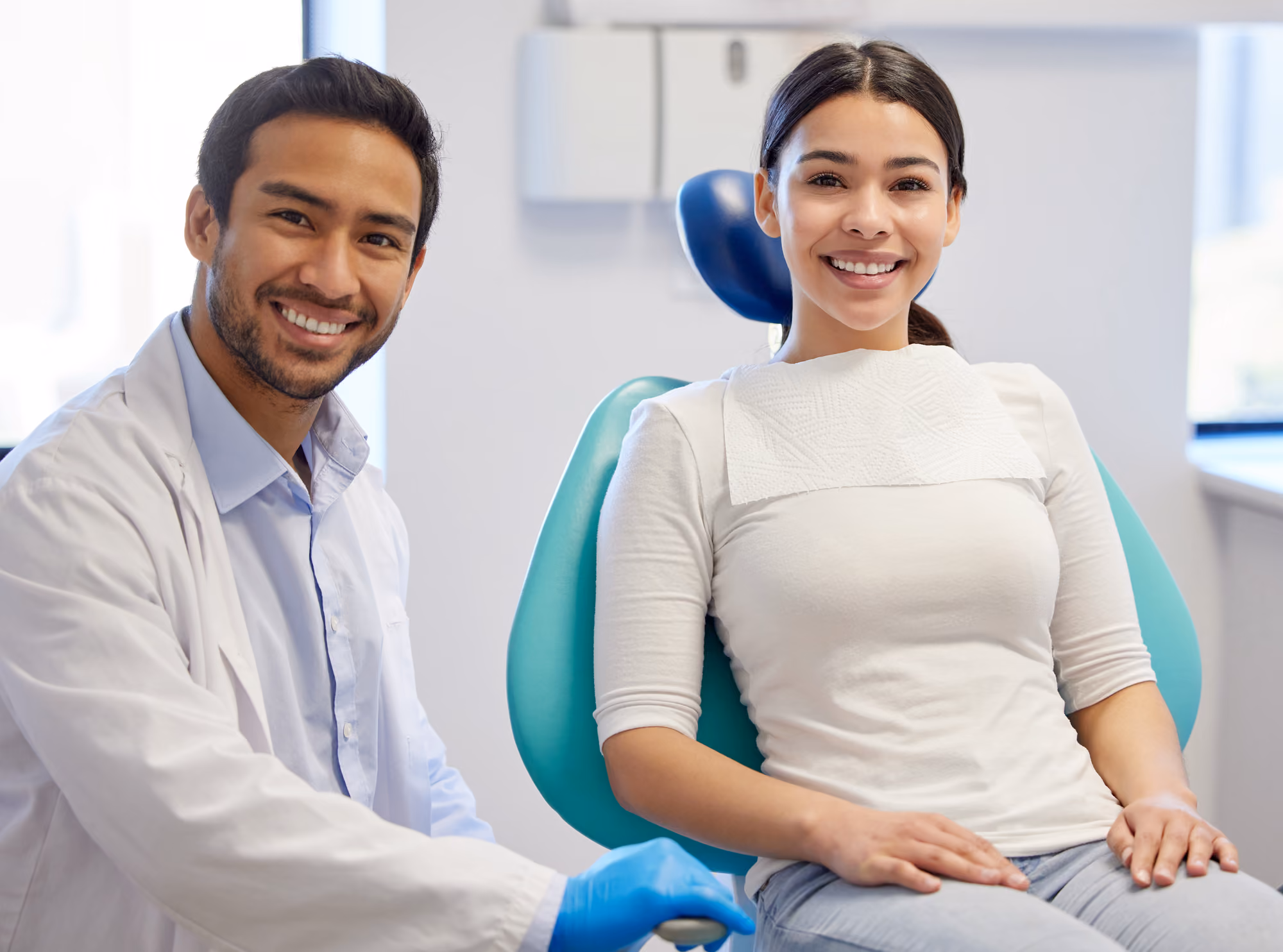 Smiling dentist wearing gloves next to a female patient seated in a dental chair with bib.