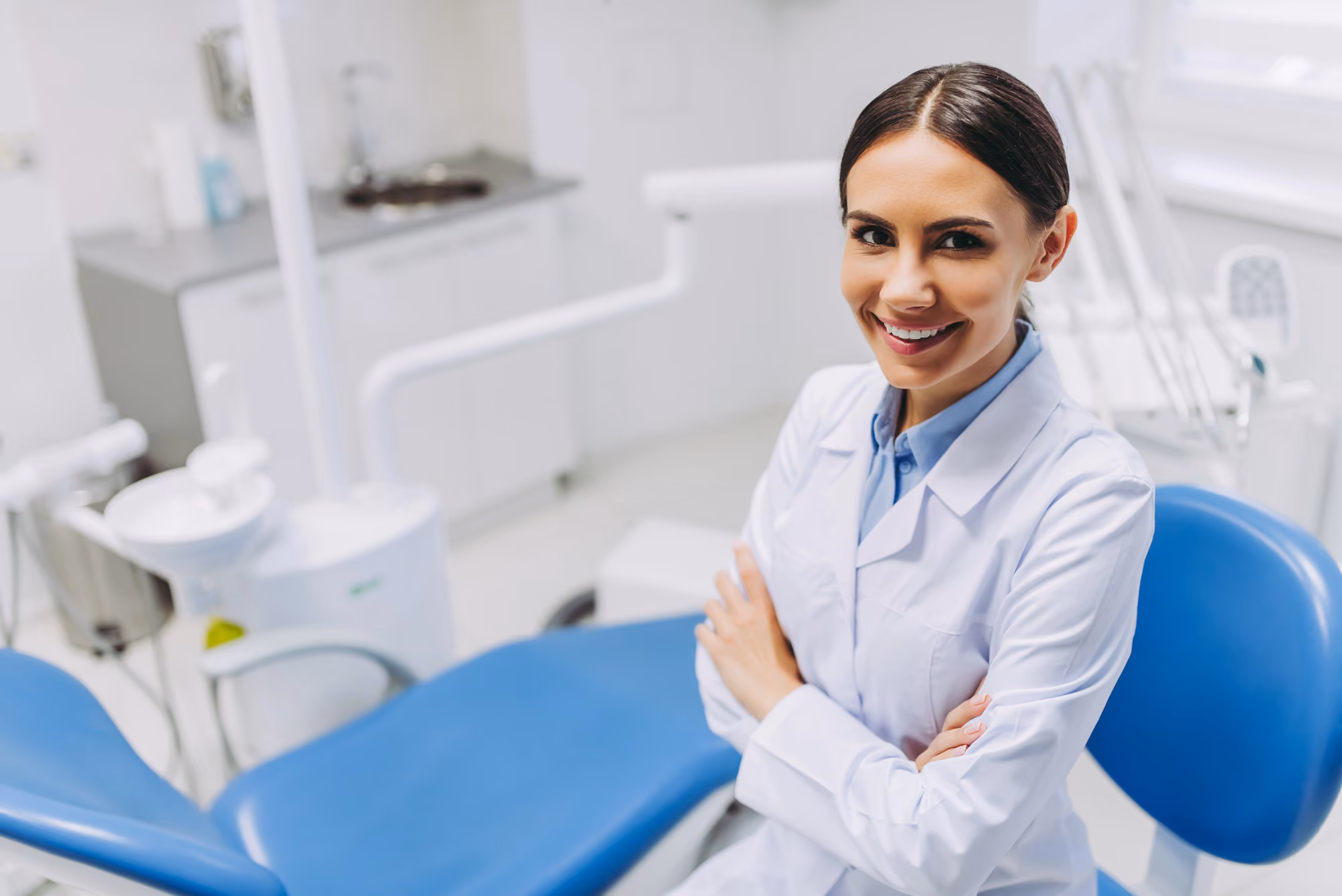 Smiling female dentist in white coat standing with arms crossed in dental clinic with blue chair.