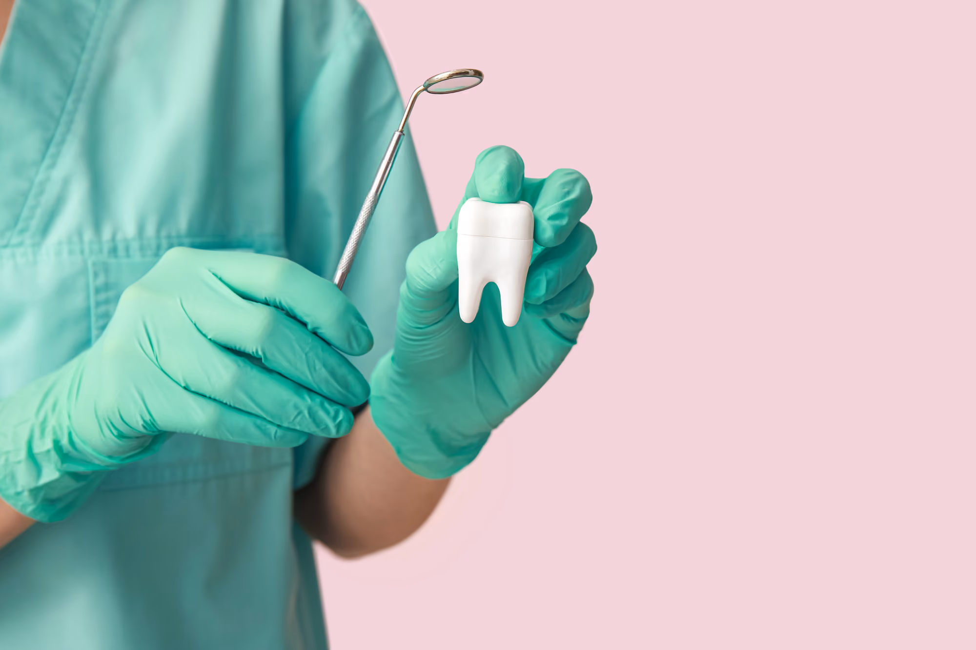 Dentist in green gloves holding a dental mirror and a large white tooth model against pink background.
