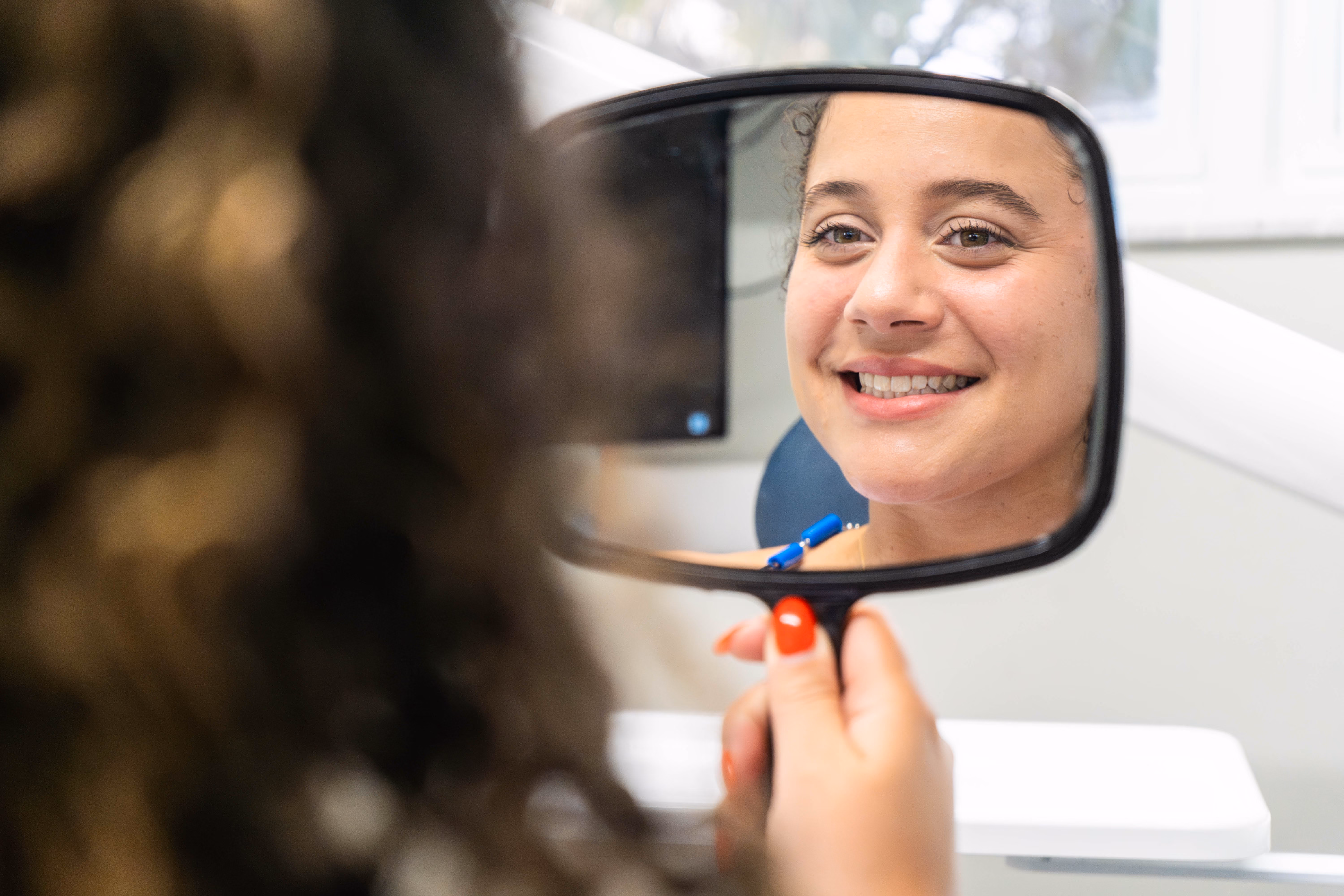Woman with curly hair smiling at her reflection in a handheld mirror after a dental cleaning.
