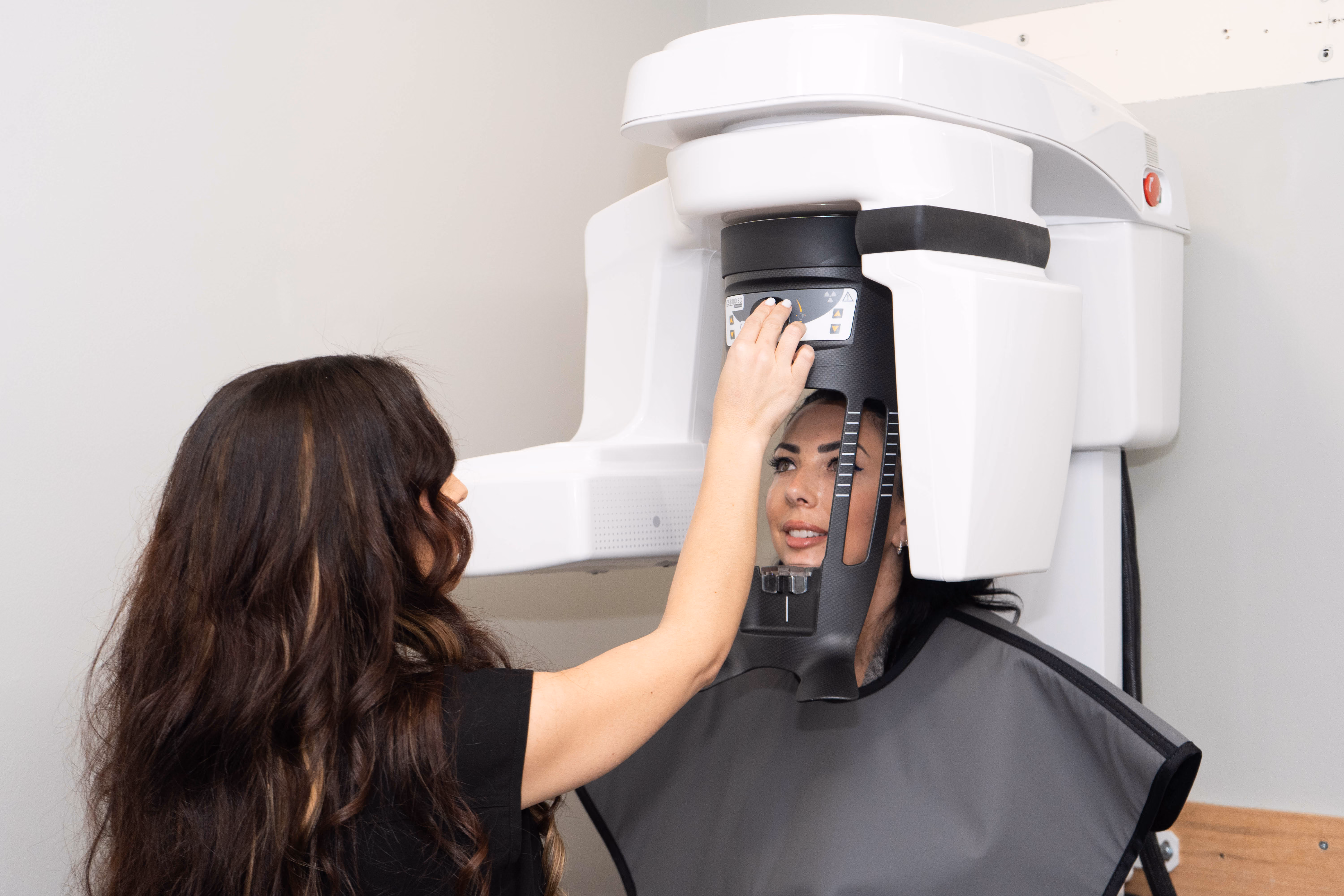 Technician adjusts a dental X-ray machine while a patient wears a protective lead apron.