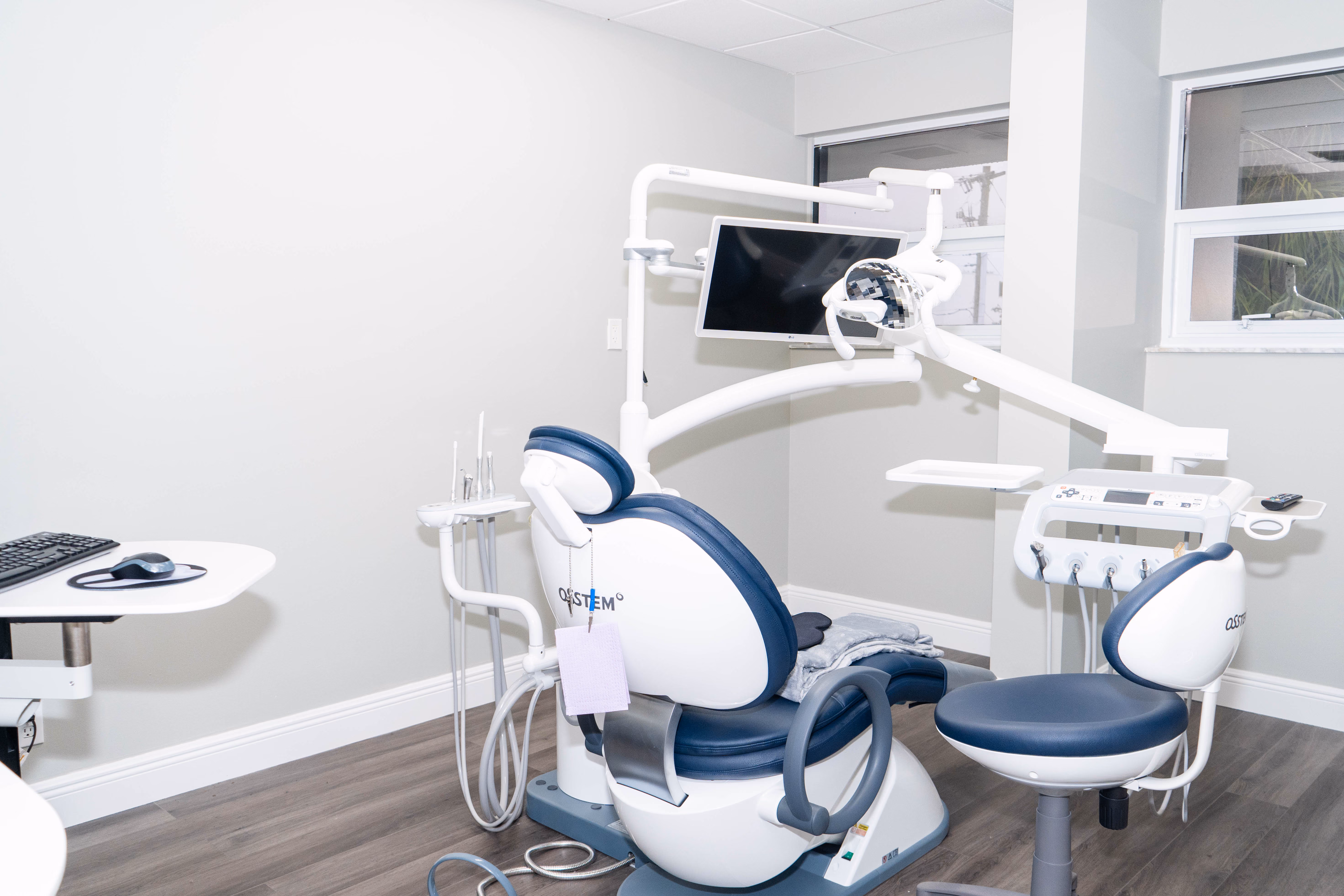 Modern dental office with blue and white dental chair, equipment, and a monitor on the wall.