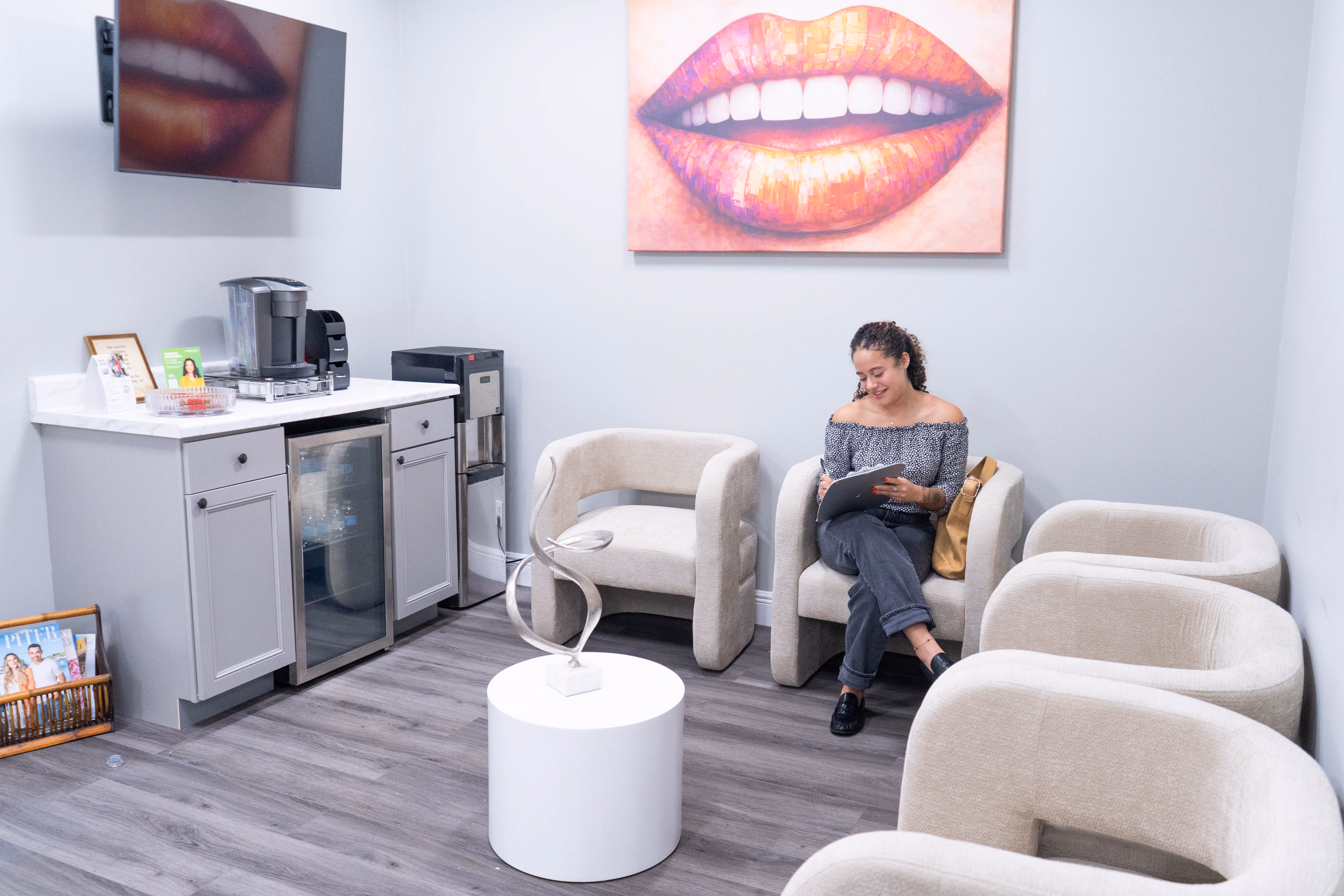 Waiting room with a woman sitting and writing, cream chairs, a round table, and a large lips painting on wall.