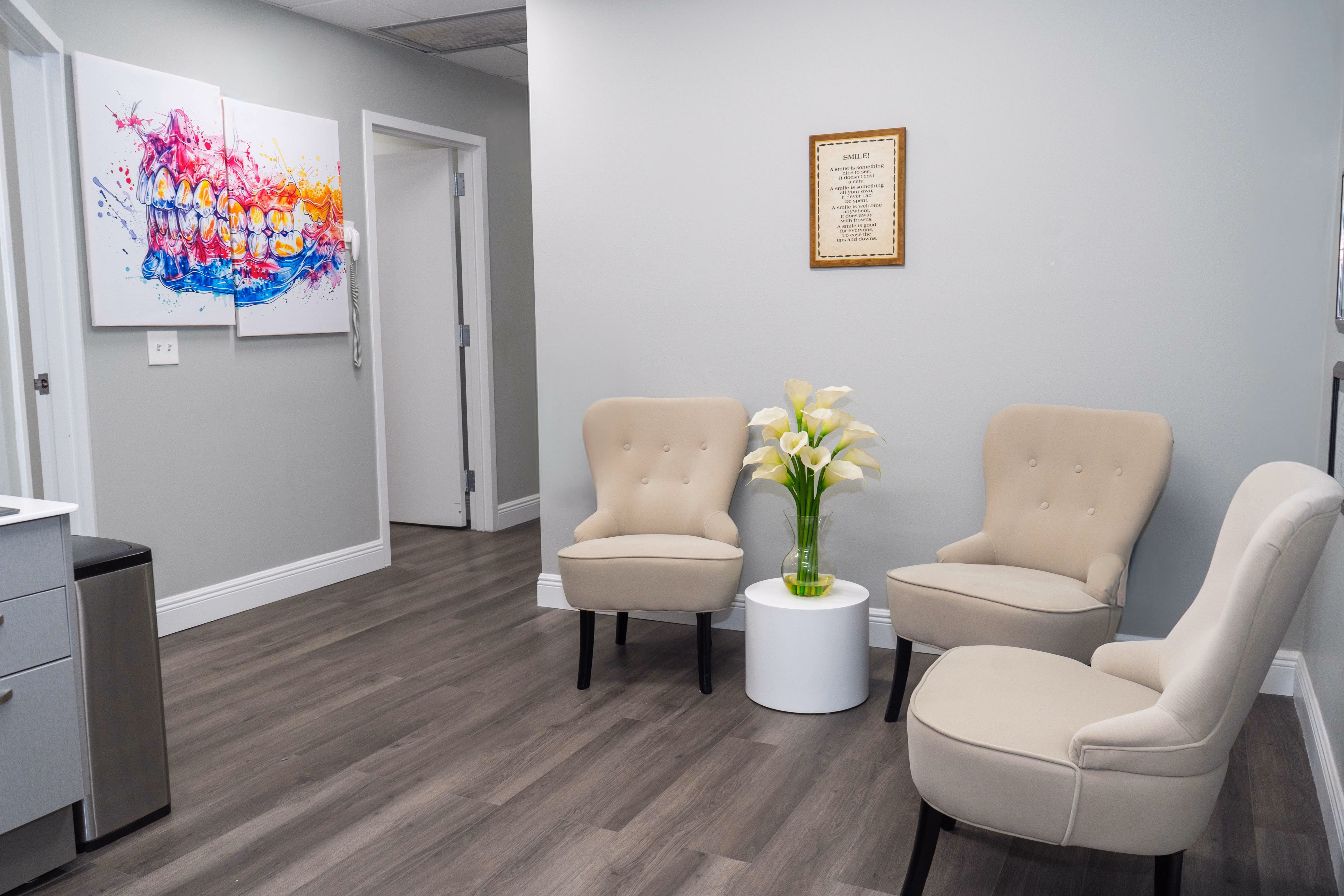 Waiting room with three beige chairs, a white side table with flowers, and colorful dental art on wall.