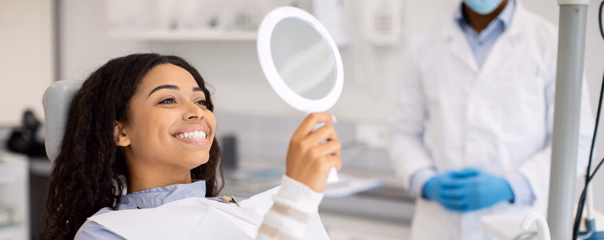 Smiling woman in dental chair holding mirror, dentist in white coat and gloves in background.