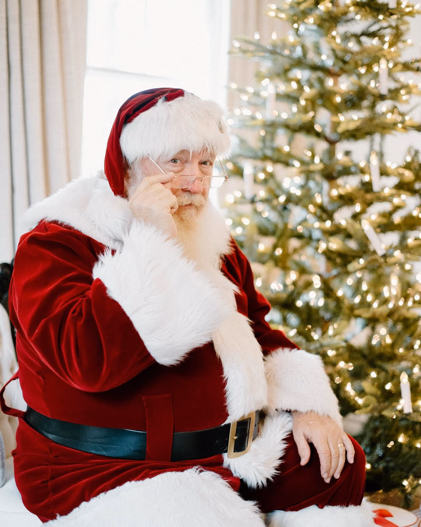 Santa Claus in red suit adjusting glasses sitting near a lit Christmas tree indoors.