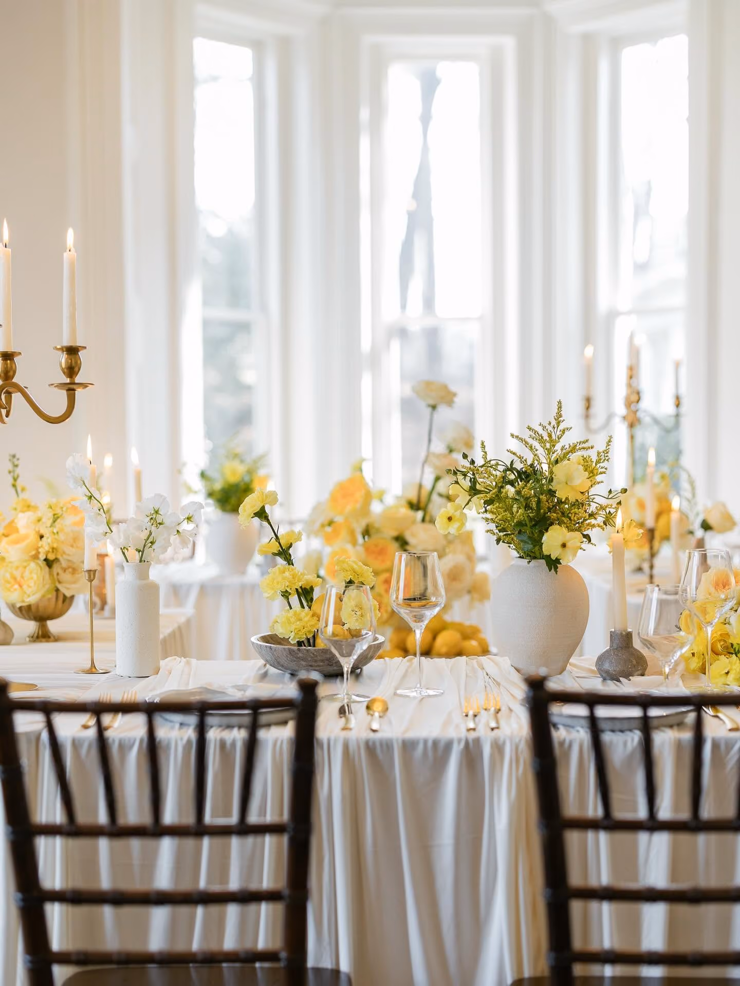 Elegant dining table with white tablecloth, yellow floral arrangements, candles, and wine glasses near bright windows.