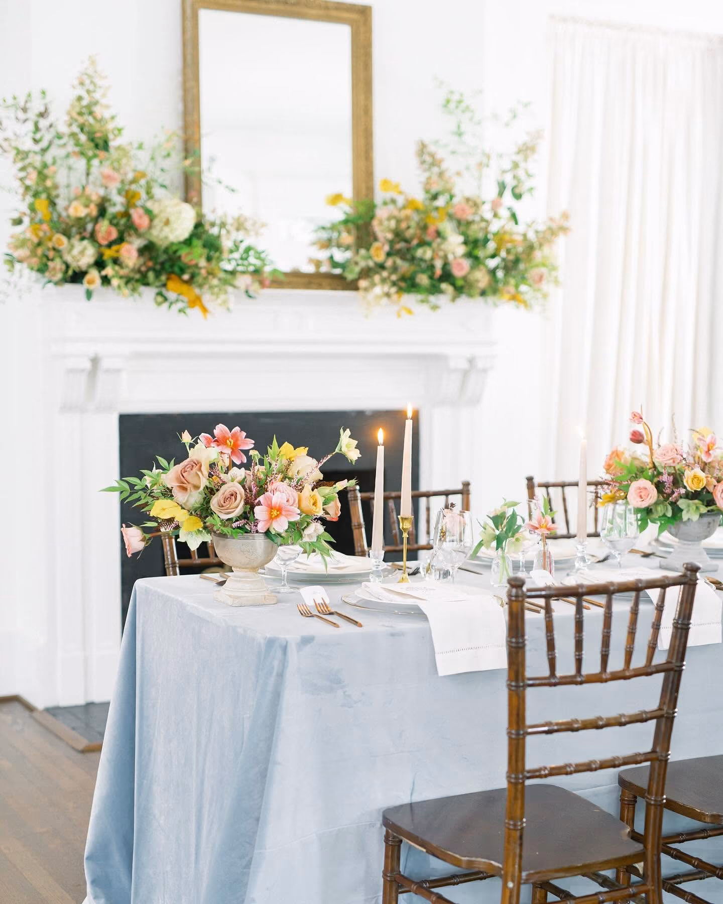 Elegant dining table with floral centerpieces, candles, and wooden chairs in a bright room with white fireplace.